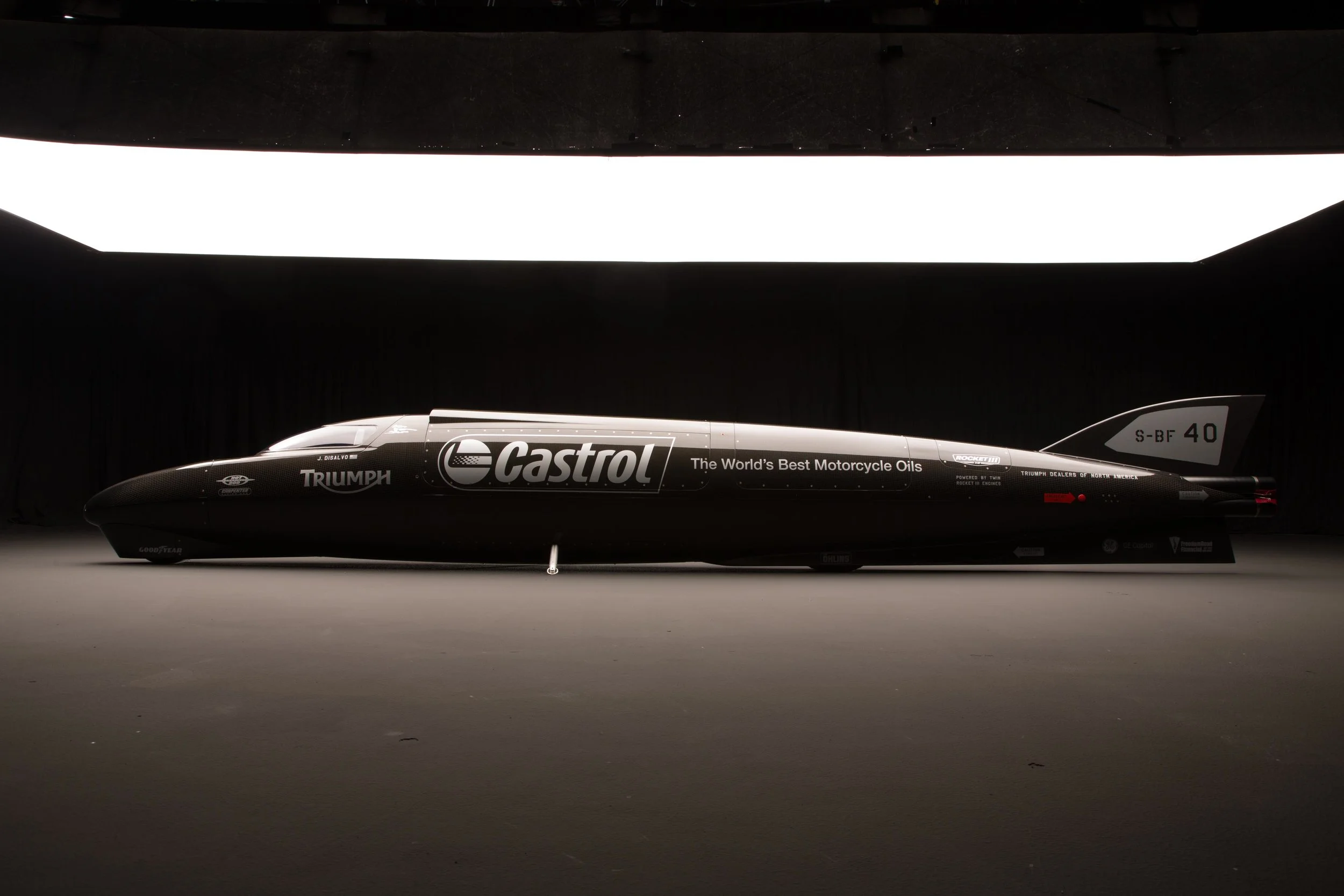 A sleek black racing motorcycle with sponsor logos including Castrol, Triumph, and Goodyear, displayed in a dark setting with a large illuminated panel in the background.