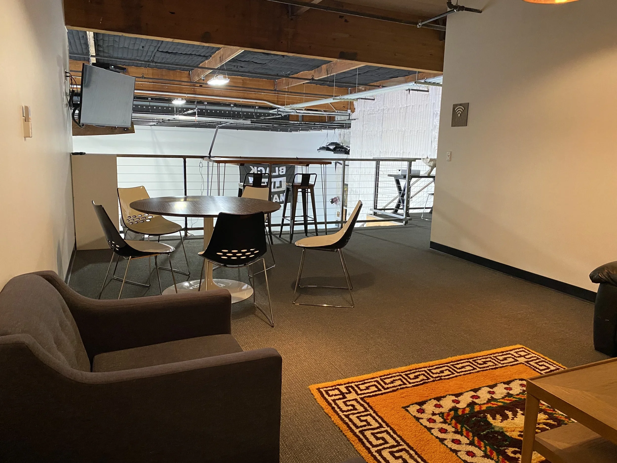 An indoor space with a sitting area, including a brown armchair and black chairs around a round table. In the background, there's a space with tables and chairs, exposed wooden beams on the ceiling, a television, and a door leading outside. A decorat