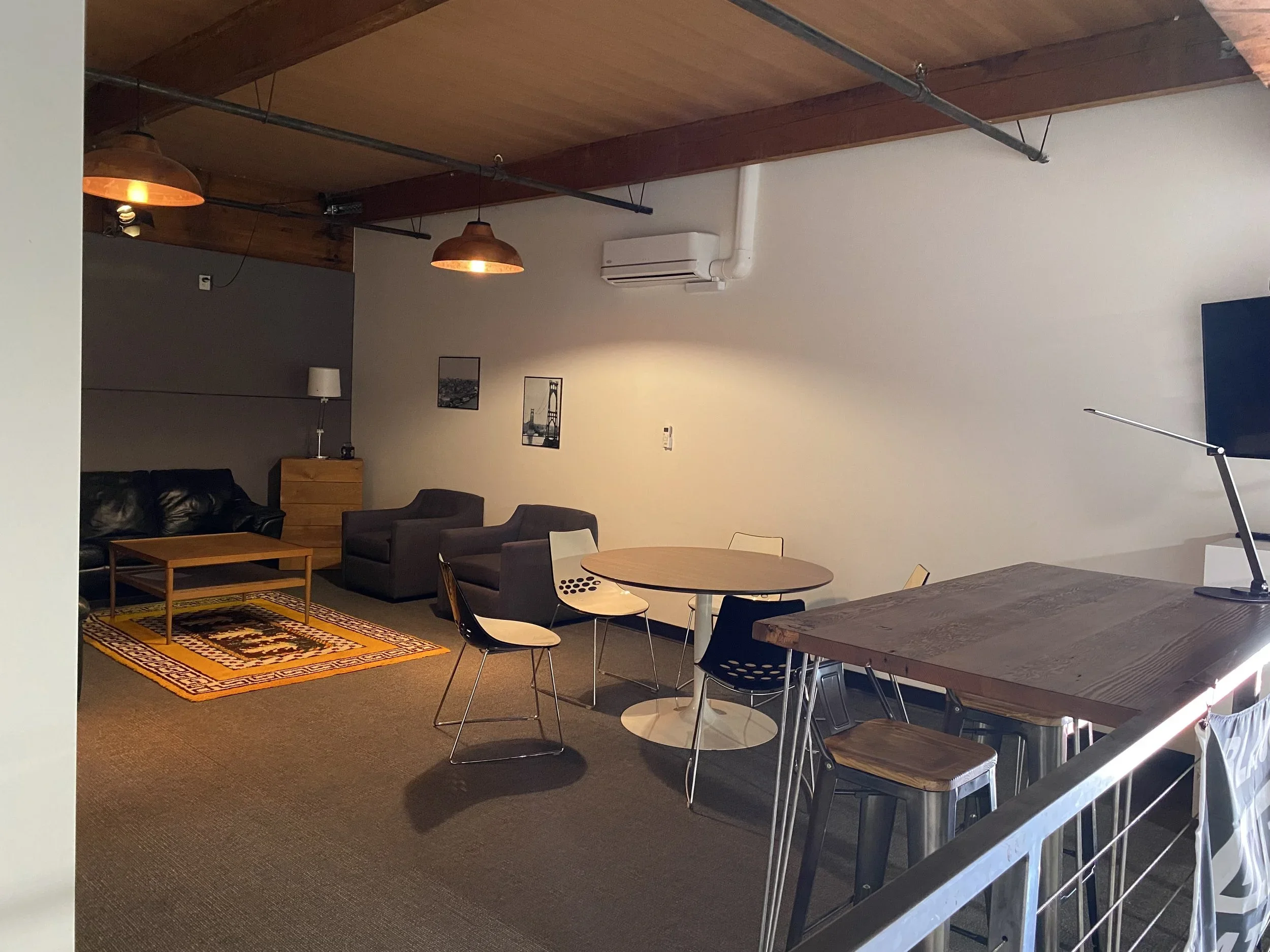 Interior view of a modern living space with wooden vaulted ceiling, hanging copper pendant lights, a gray accent wall with framed art, a black leather sofa, wooden coffee table, and a patterned rug. Adjacent is a dining area with a round wooden table