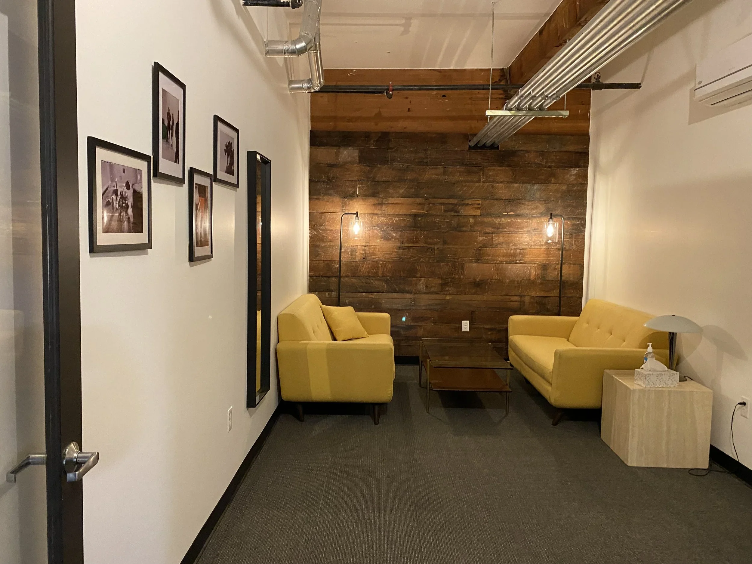 Interior view of a cozy sitting area with yellow armchairs, a side table, wooden wall, framed photos on white wall, and warm lighting.