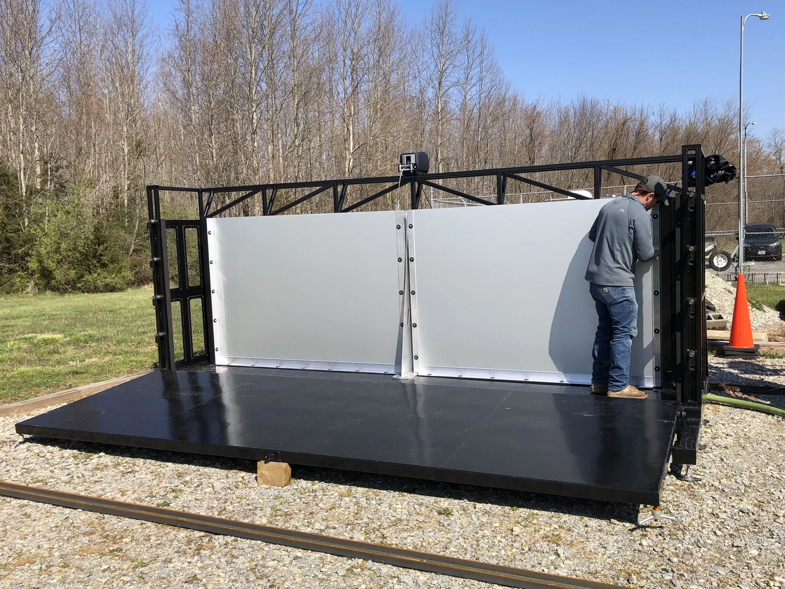 A person in a gray jacket and cap is working on a large whiteboard or screen mounted on a black platform outdoors. The sky is clear, and leafless trees are in the background.