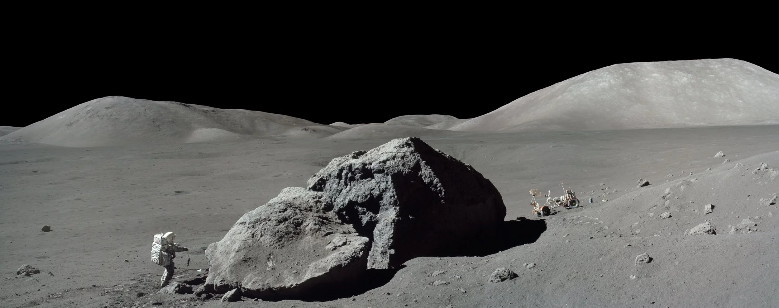 An astronaut on the moon's surface near a large rock, with lunar terrain and hills in the background, and a lunar rover in the distance.