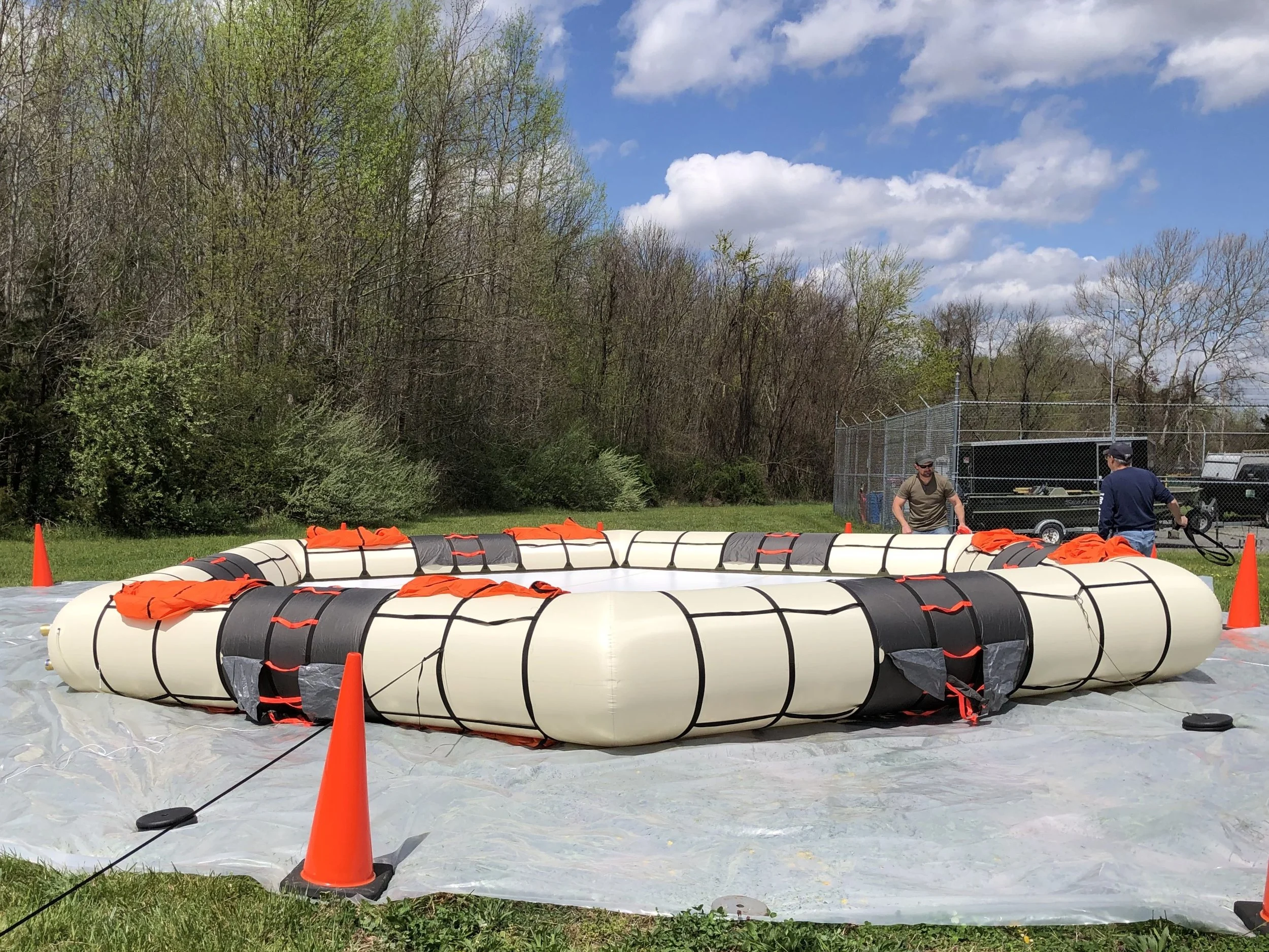 Large inflatable pool with orange life jackets inside, surrounded by orange traffic cones on a grassy area. Two men are standing nearby, and there is a wooded area with trees and a fence in the background.