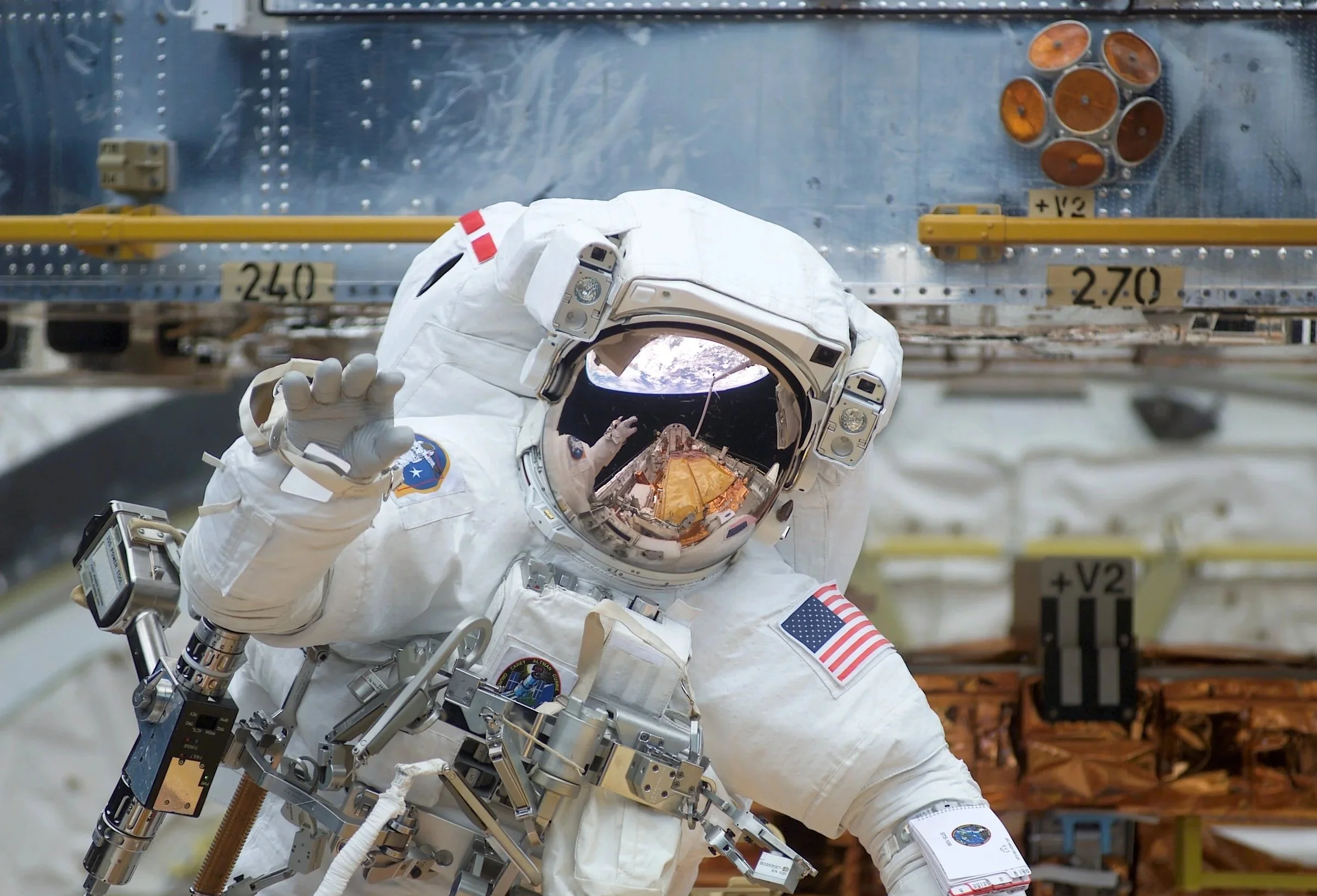 An astronaut in a space suit floating near a spacecraft, with a view of Earth reflected in the helmet's visor.