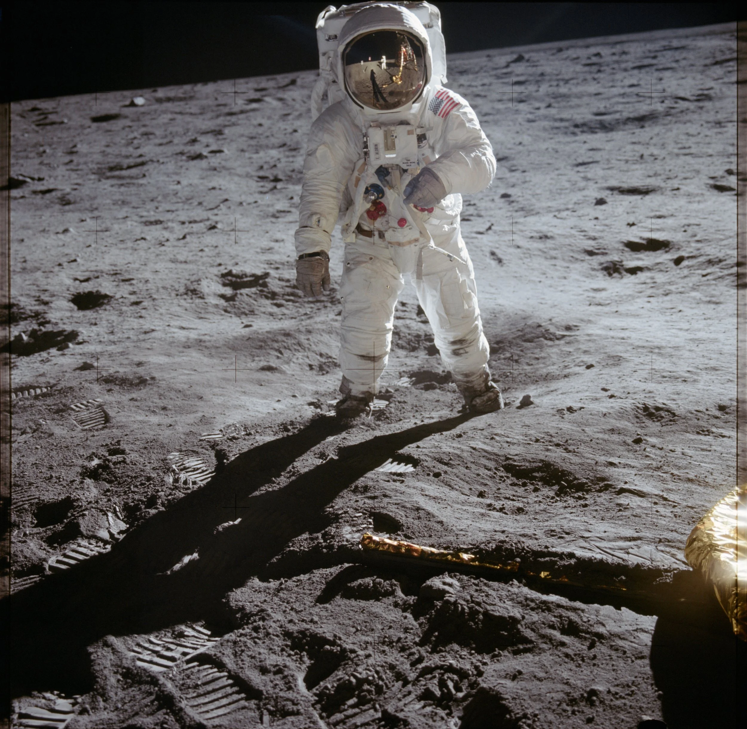 An astronaut on the moon in a white space suit with an American flag patch points at the moon's surface, which has tracks from a lunar rover. The reflection of the astronaut's helmet shows the lunar module and the Earth in the sky.