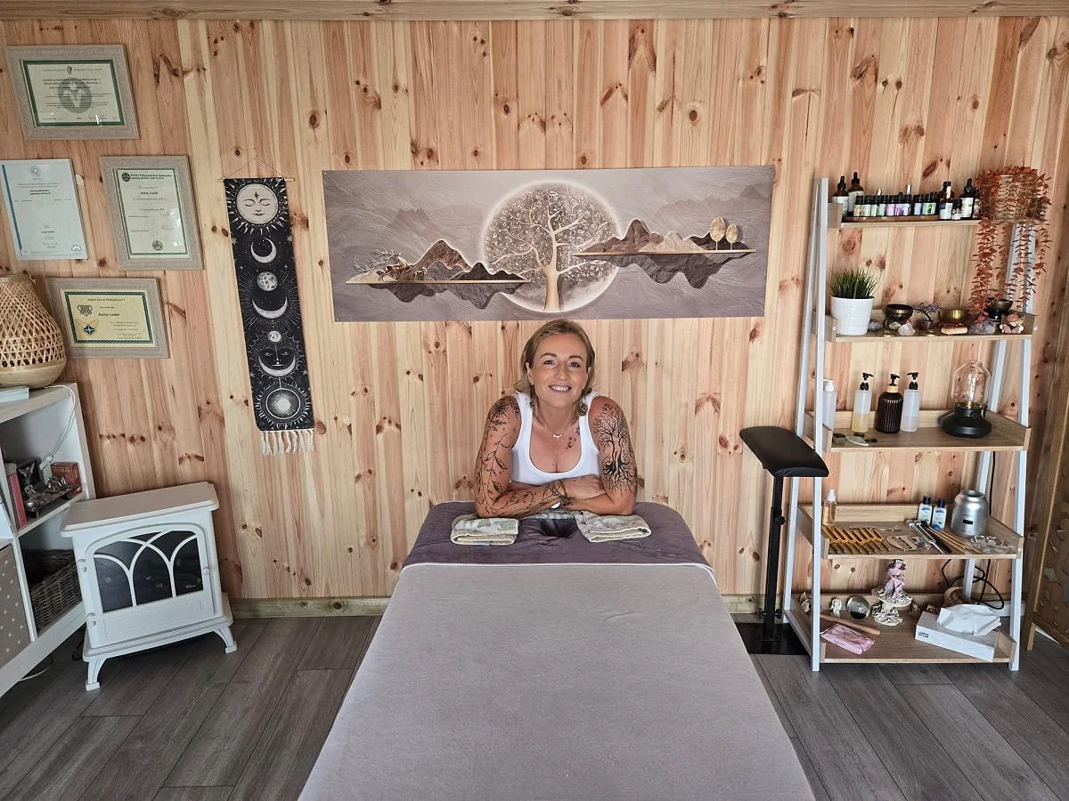 A woman with tattoos smiling while lying on her stomach on a massage table in a wellness room decorated with wood paneling, certificates on the wall, and shelves with essential oils, plants, and wellness items.