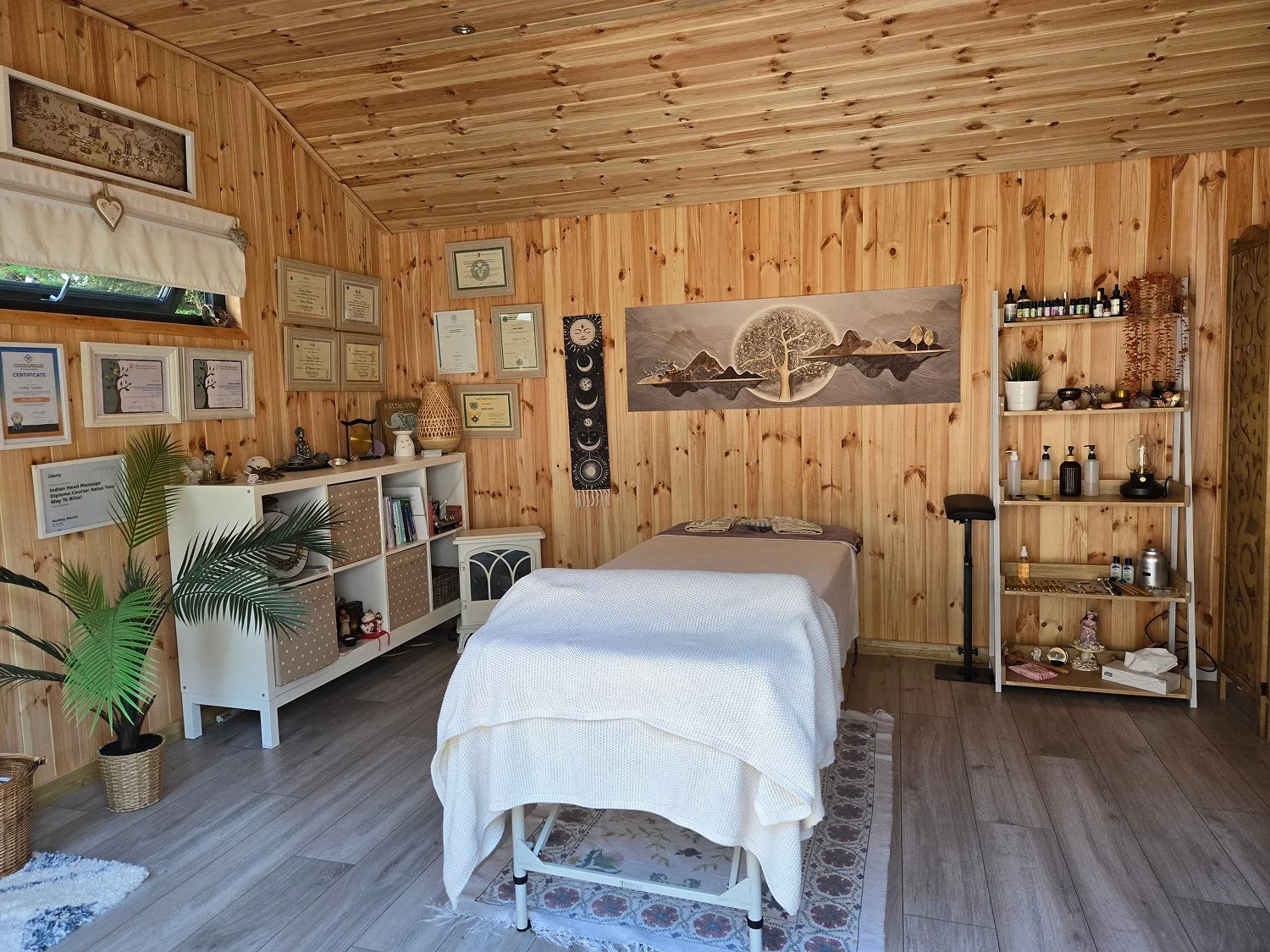 A wellness or massage room with wooden walls and ceiling, featuring a massage table covered with a white sheet, surrounded by certificates and decor, including plants, shelves with bottles and bowls, and a large wall art piece depicting a tree, mountains, and a sun.