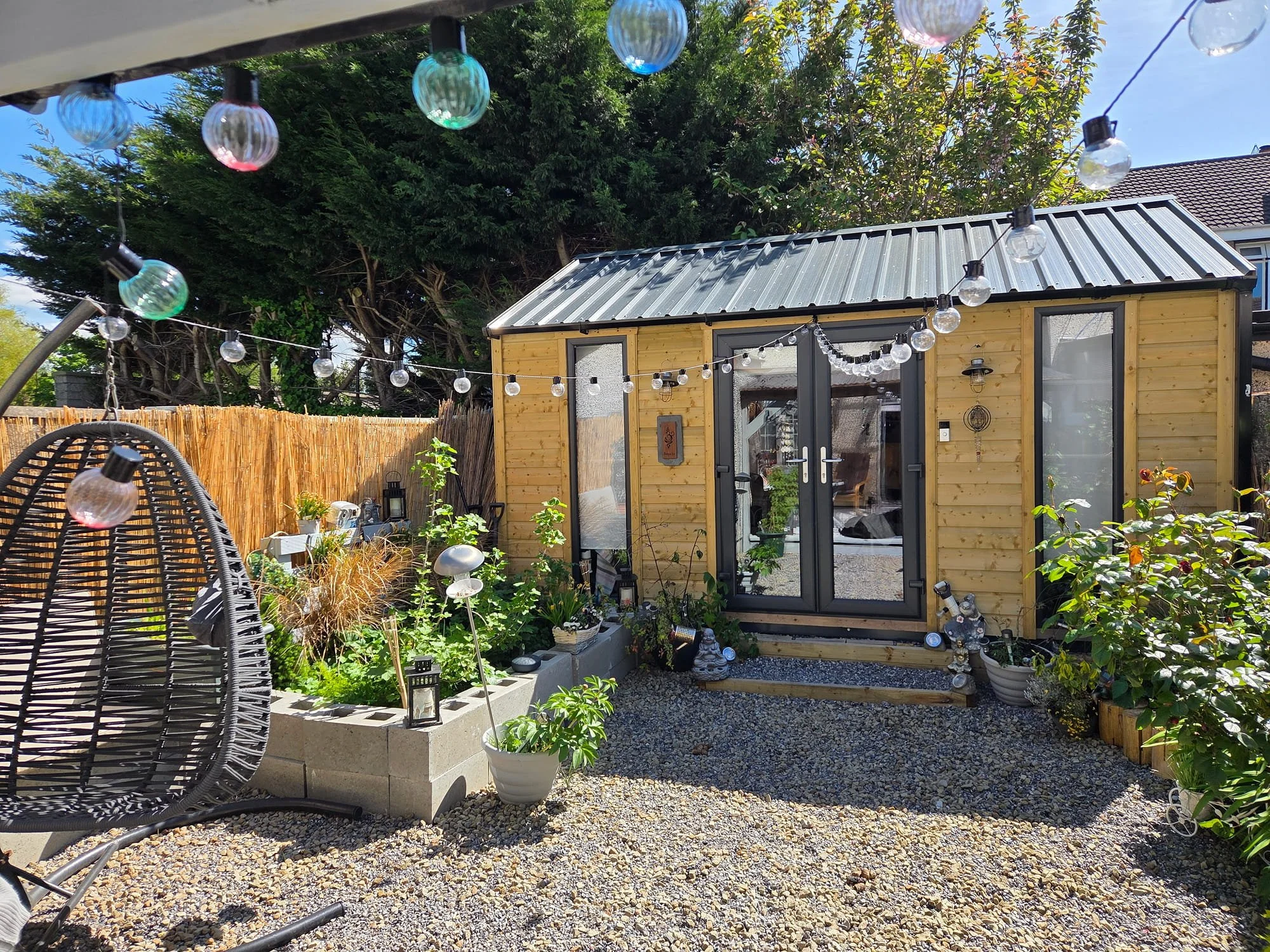 A backyard patio featuring a small wooden shed with a metal roof, string lights hanging overhead, various potted plants, gravel ground, a wicker egg-shaped chair, and lush green trees in the background.