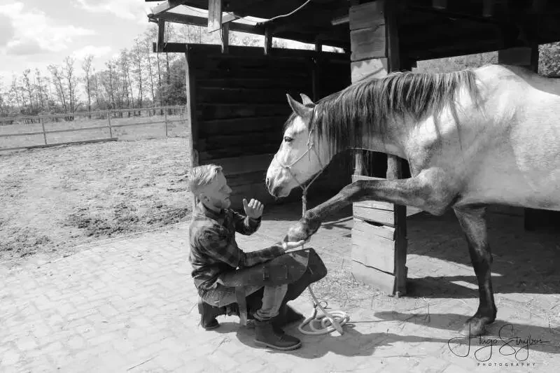 A young boy kneeling and holding a horse's hoof in a stable yard