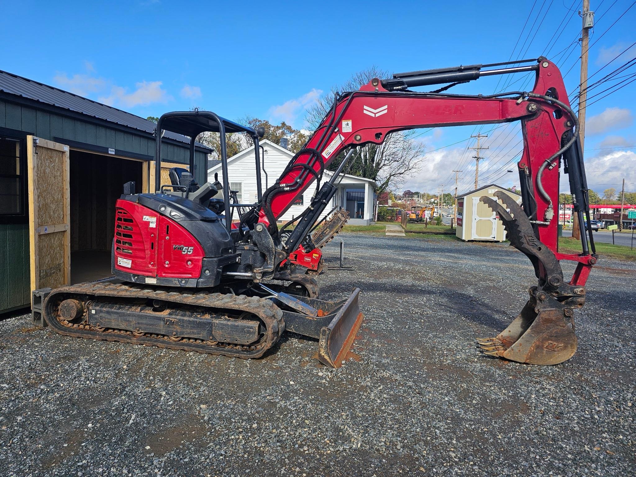 A red mini excavator with black tracks parked outside on gravel, next to a building under construction, with a white house and power lines in the background.