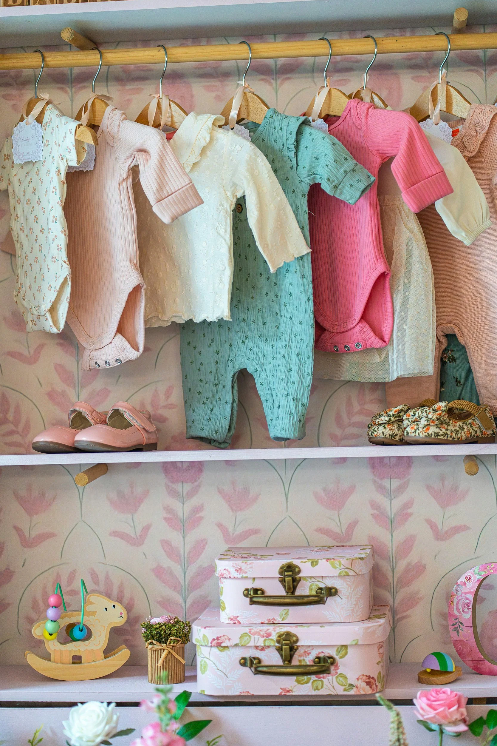 Baby clothes hanging on a rack, including various pastel-colored onesies and pajamas, with pink floral wallpaper in the background. Below are decorative items like a wooden rocking horse, small potted plants, and pastel-colored storage boxes.