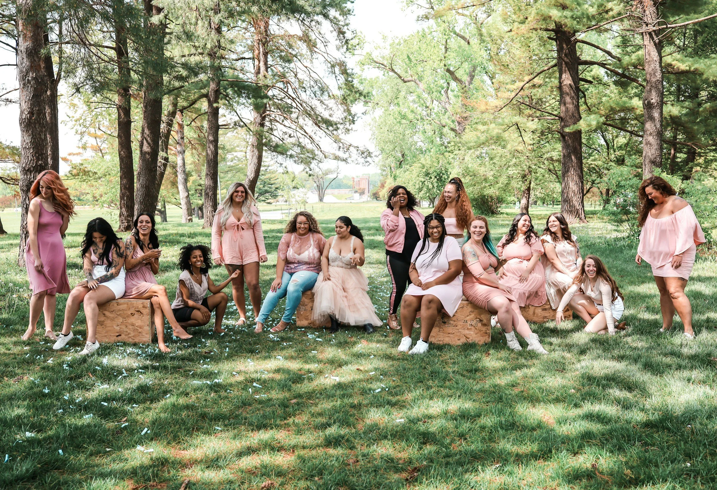 Group of diverse women in pink dresses and casual outfits smiling and gathering in a wooded park, showcasing the Divinity Doula postpartum doula and nanny‑educator team in Greenville SC.