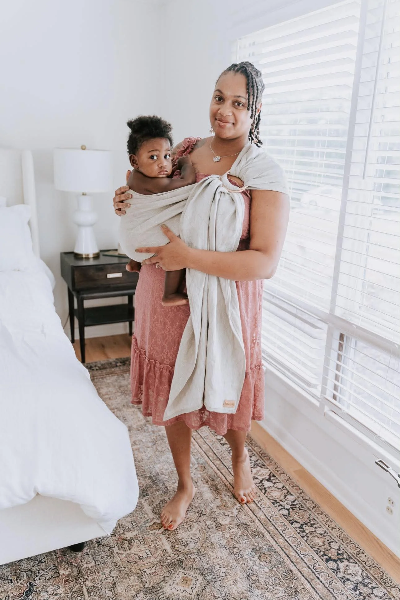 Greenville SC postpartum doula and Divinity Doula founder Jernae Webb holding her newborn in a bright bedroom with white walls, large windows, and a patterned rug.