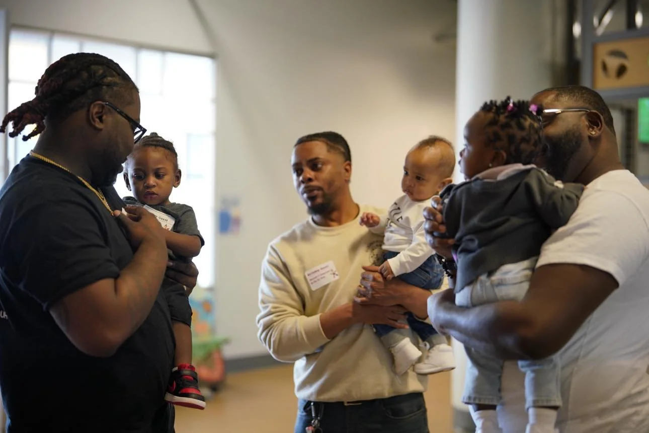 Men holding their children and connecting in a bright indoor space, representing community support for new fathers in the postpartum period.