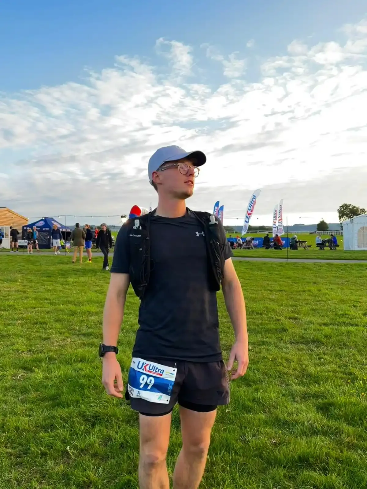 A young man wearing a black athletic outfit, glasses, a white cap, and a smartwatch stands on a grassy field during a race event. He has a race bib with the number 99. In the background, there are tents, flags, and other people, with a partly cloudy sky above.