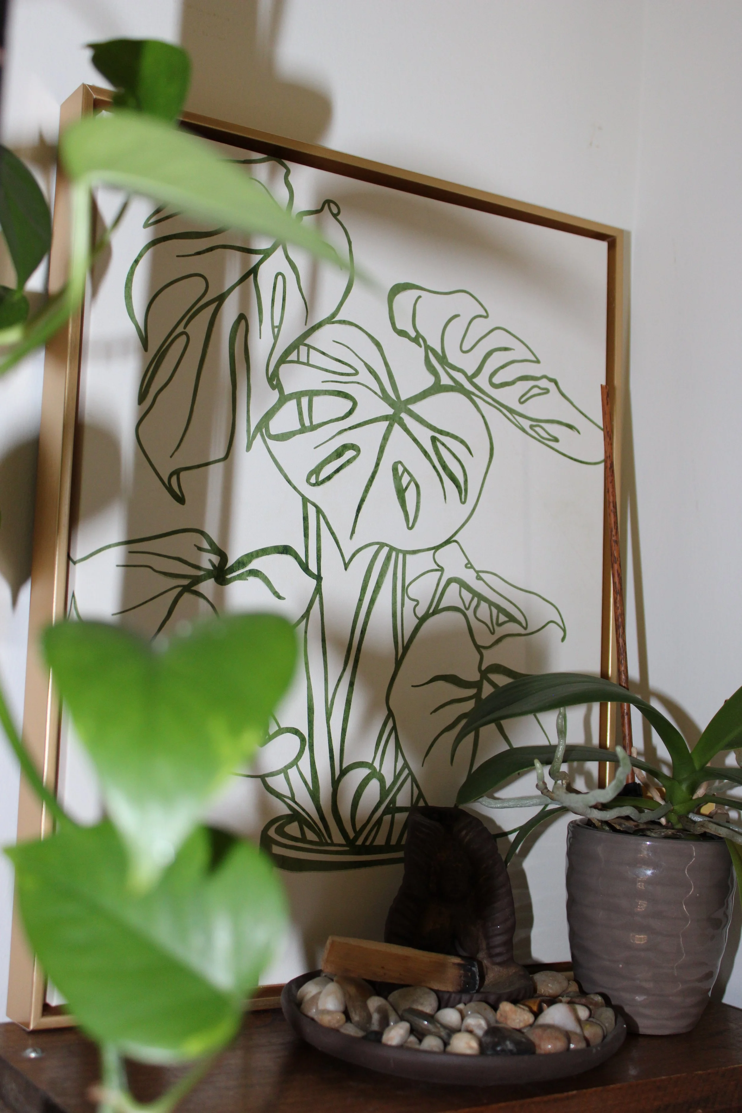 A corner of a room with houseplants and a decorative black tray filled with smooth stones and a wooden object, with a leafy plant in the foreground and a framed line drawing of tropical leaves on the wall.