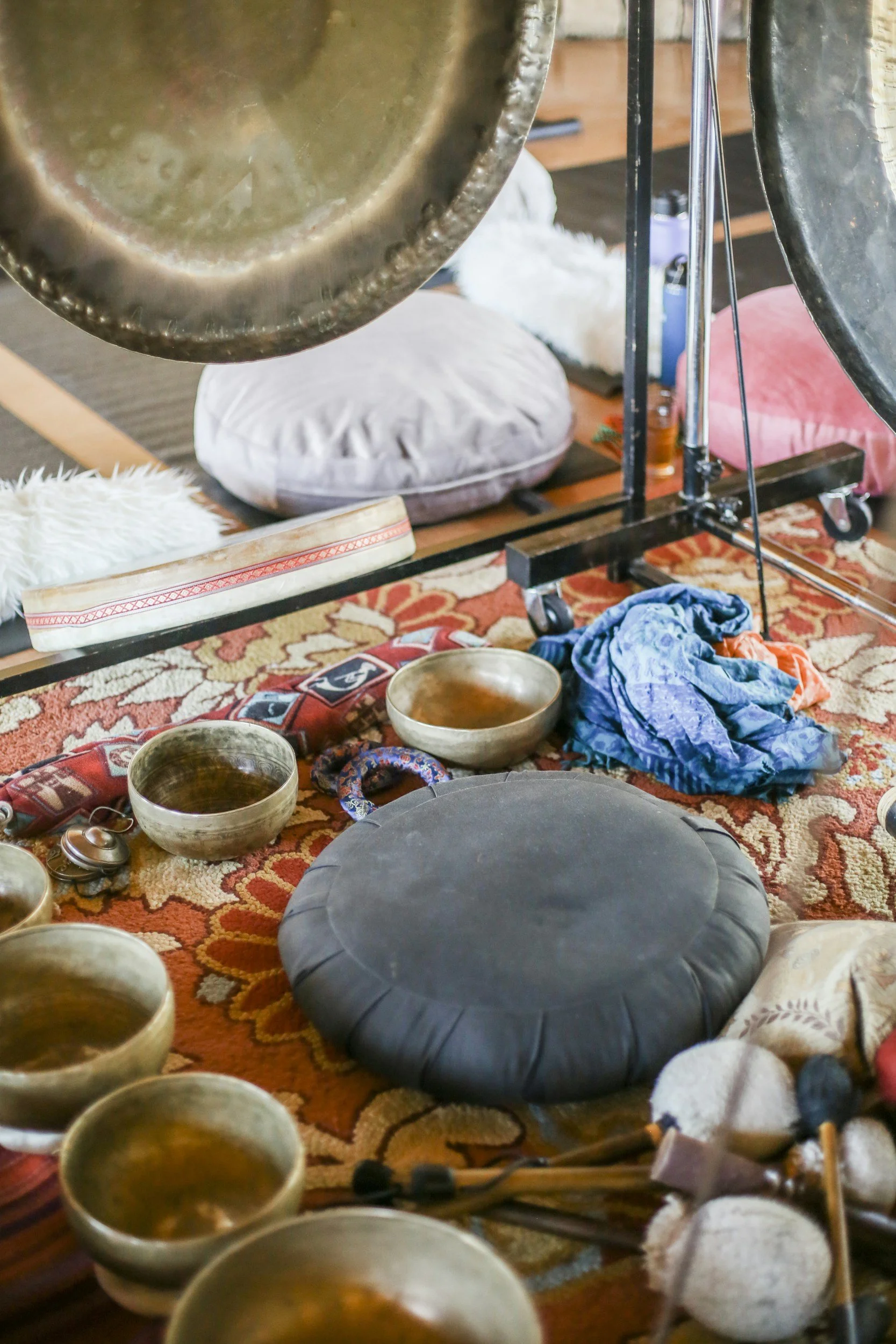 A collection of Tibetan singing bowls, cushions, and drums arranged on a decorative rug, with percussion mallets nearby.