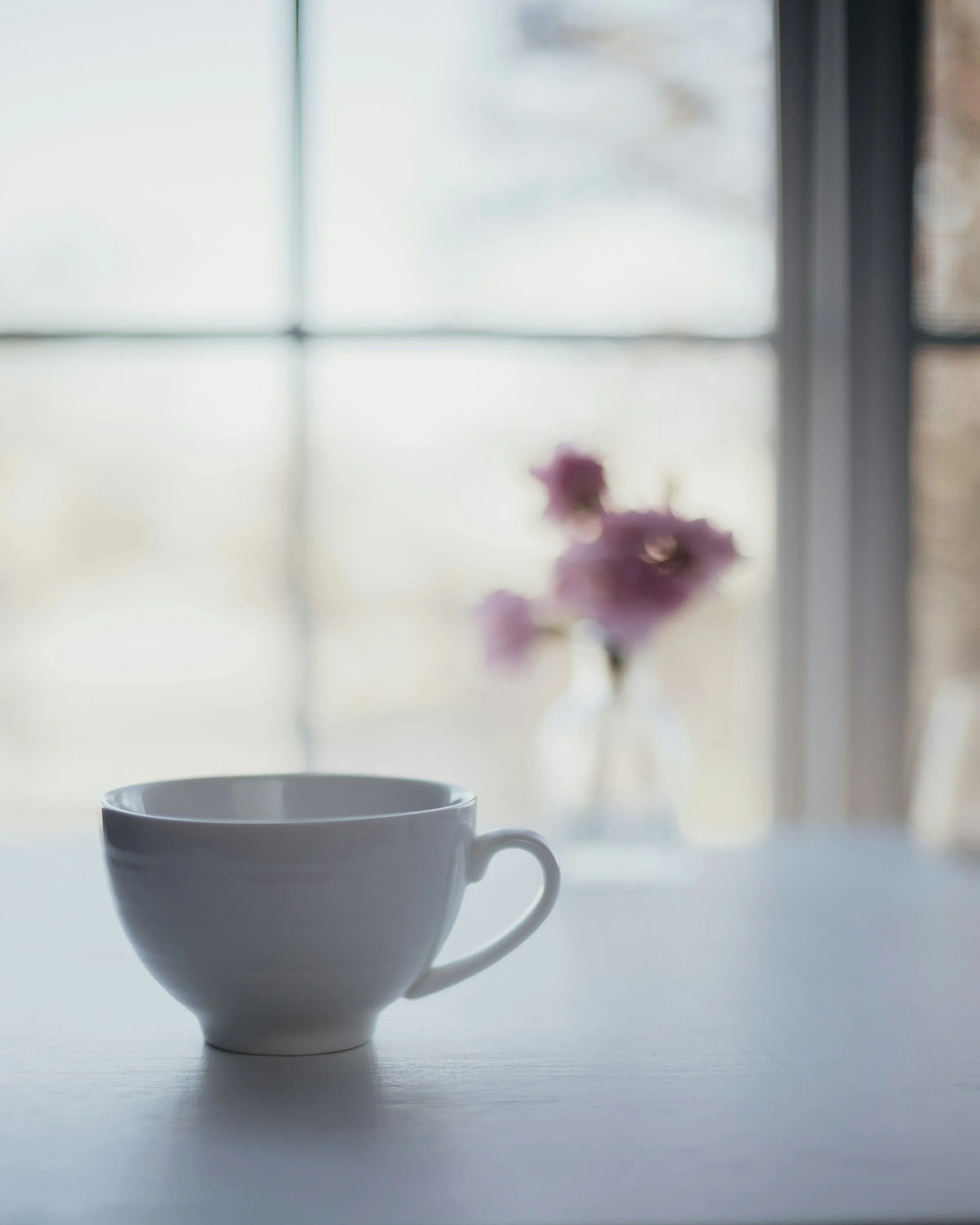 A white ceramic cup with a handle on the right side sitting on a white surface, with a blurred window and a flower in a vase in the background.