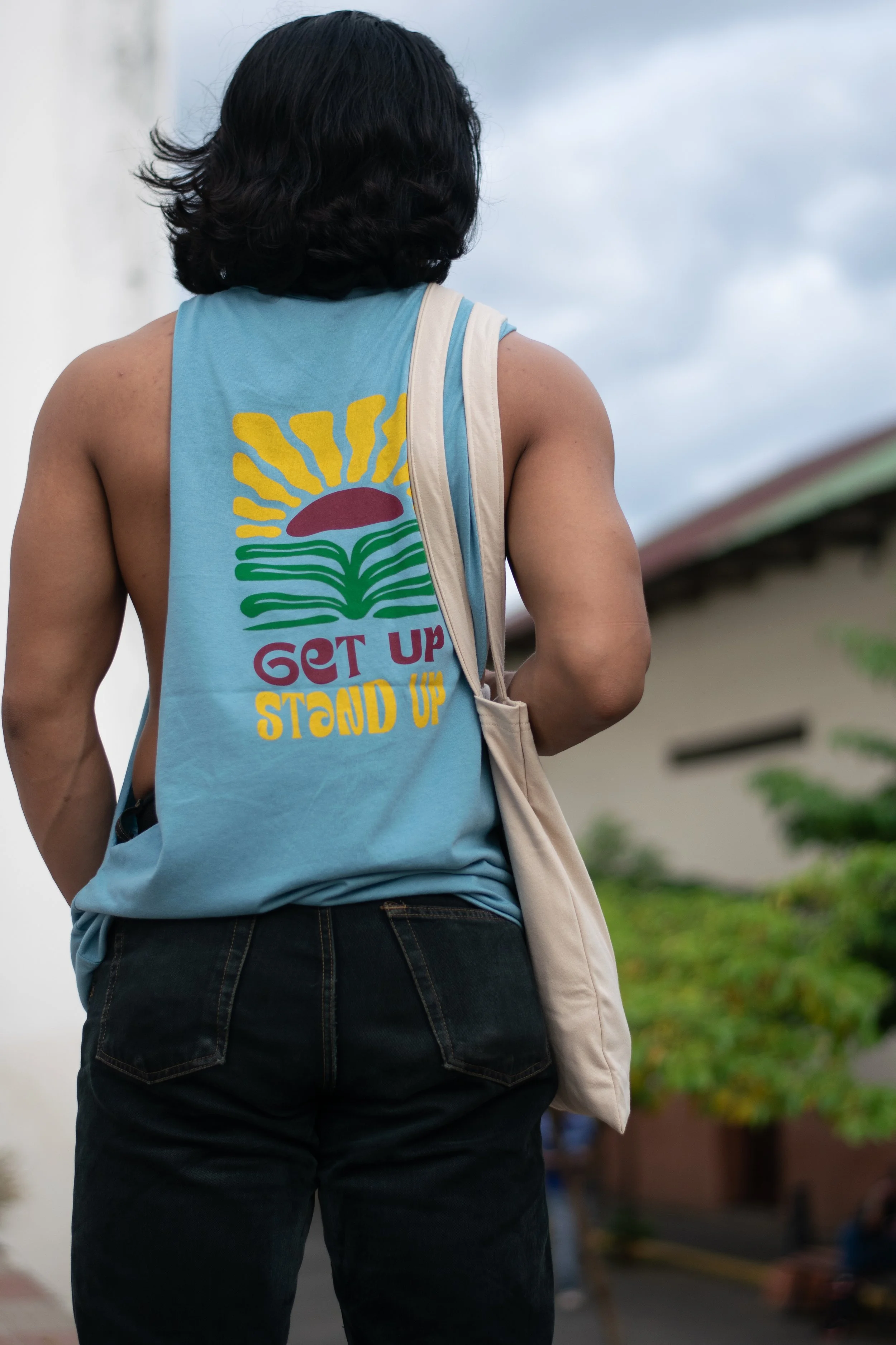 Back view of a person with medium hair wearing a sleeveless blue T-shirt with a colorful sun and plant design, carrying a beige bag, walking outdoors in a residential area during daytime.