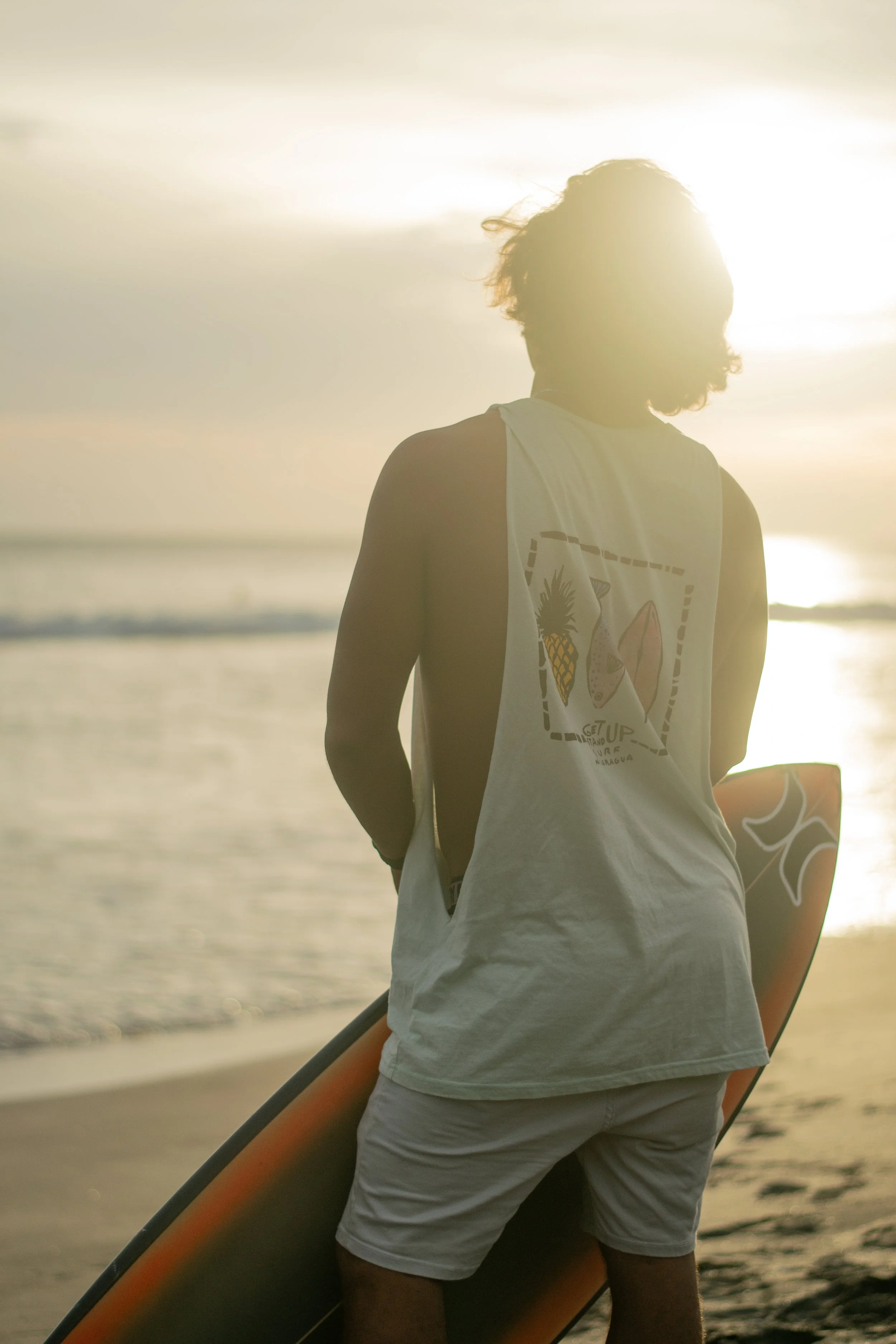 Person standing on the beach holding a surfboard during sunset.