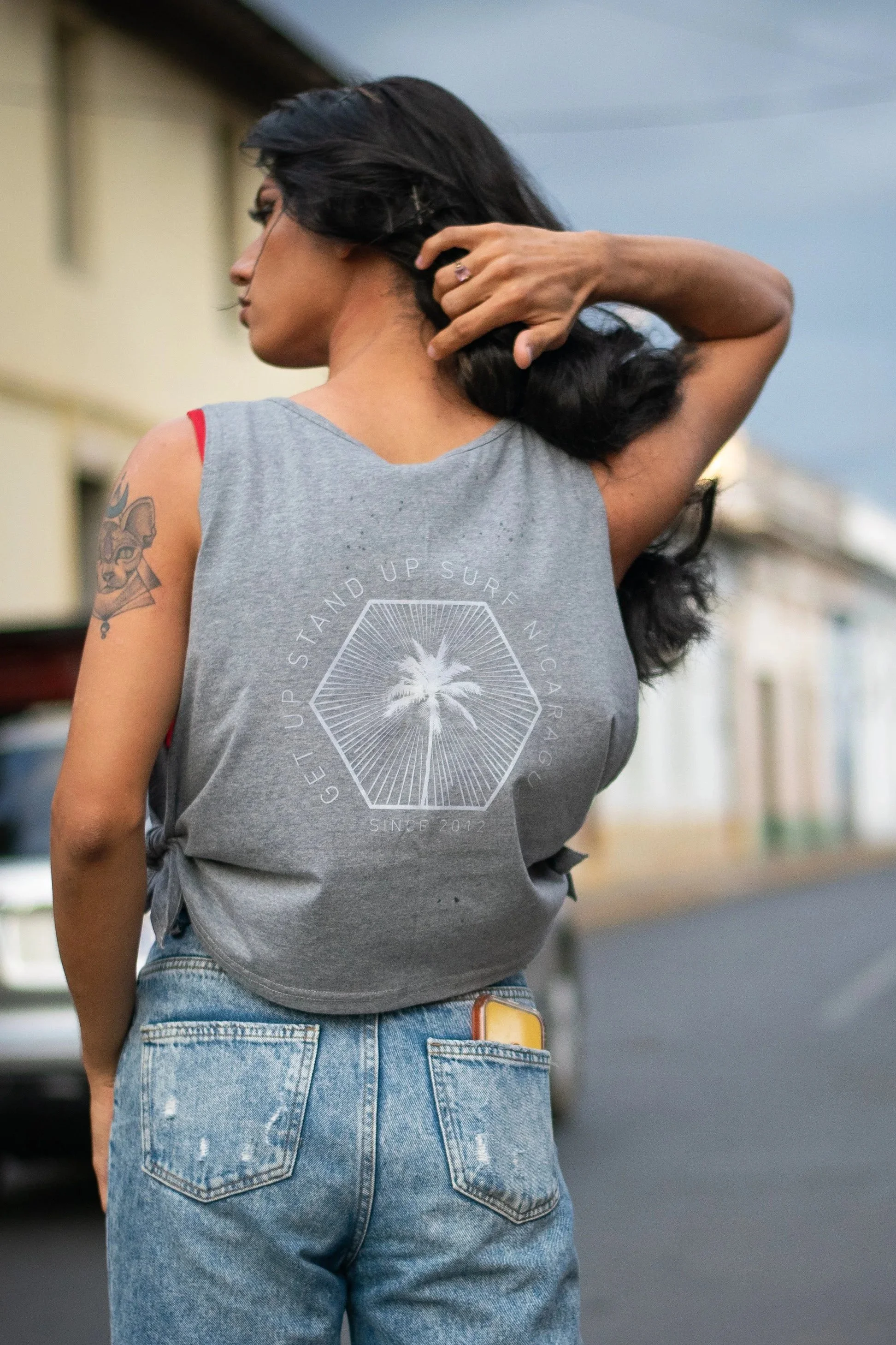 A woman with dark hair and a tattoo on her upper arm, wearing a gray sleeveless shirt and jeans, standing outdoors near a building with her hand behind her head.
