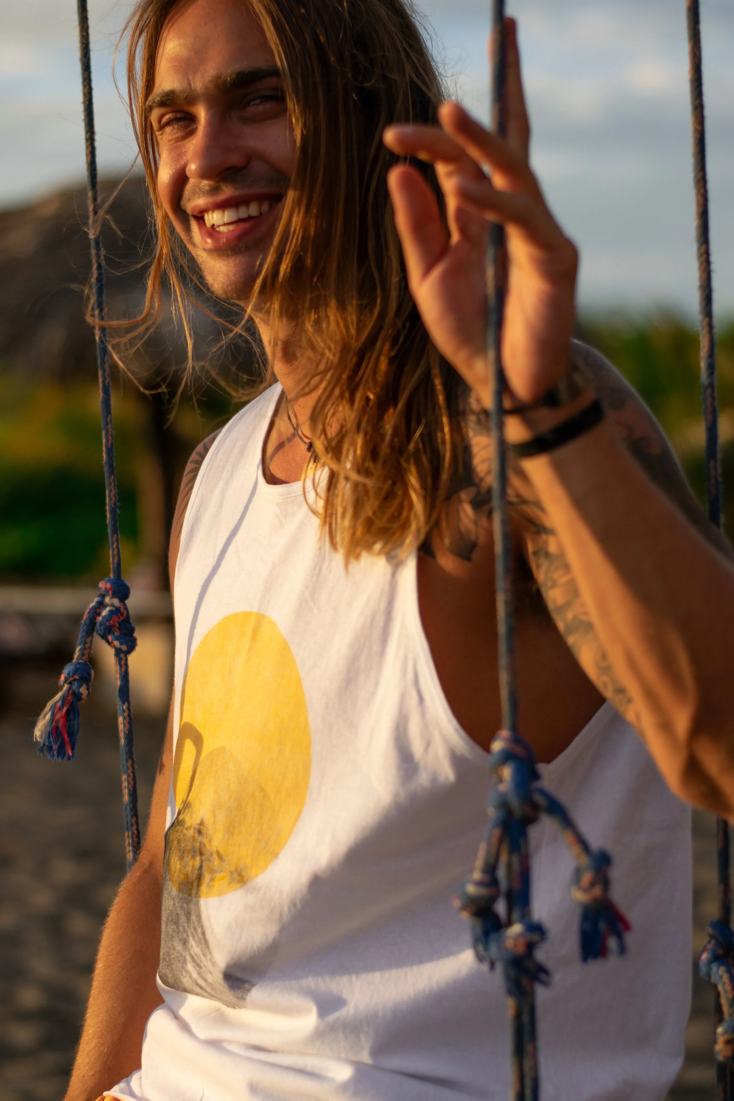 Close-up of a smiling man with long hair, wearing a white sleeveless shirt with a yellow sun graphic, standing outdoors near a swing, during sunset.