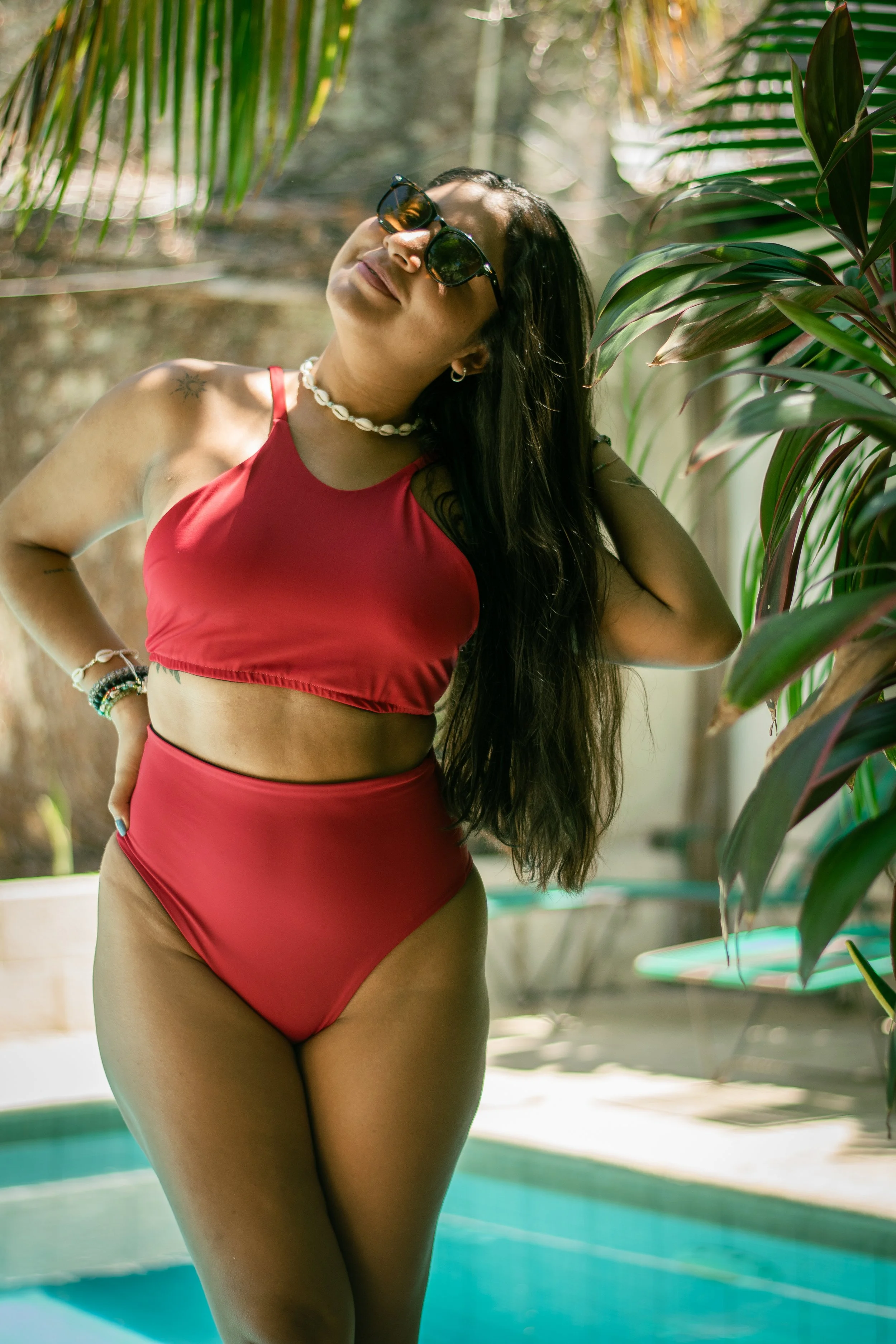 Woman with long dark hair wearing sunglasses, red bikini top, and shell necklace, standing outdoors near a pool with greenery in the background.