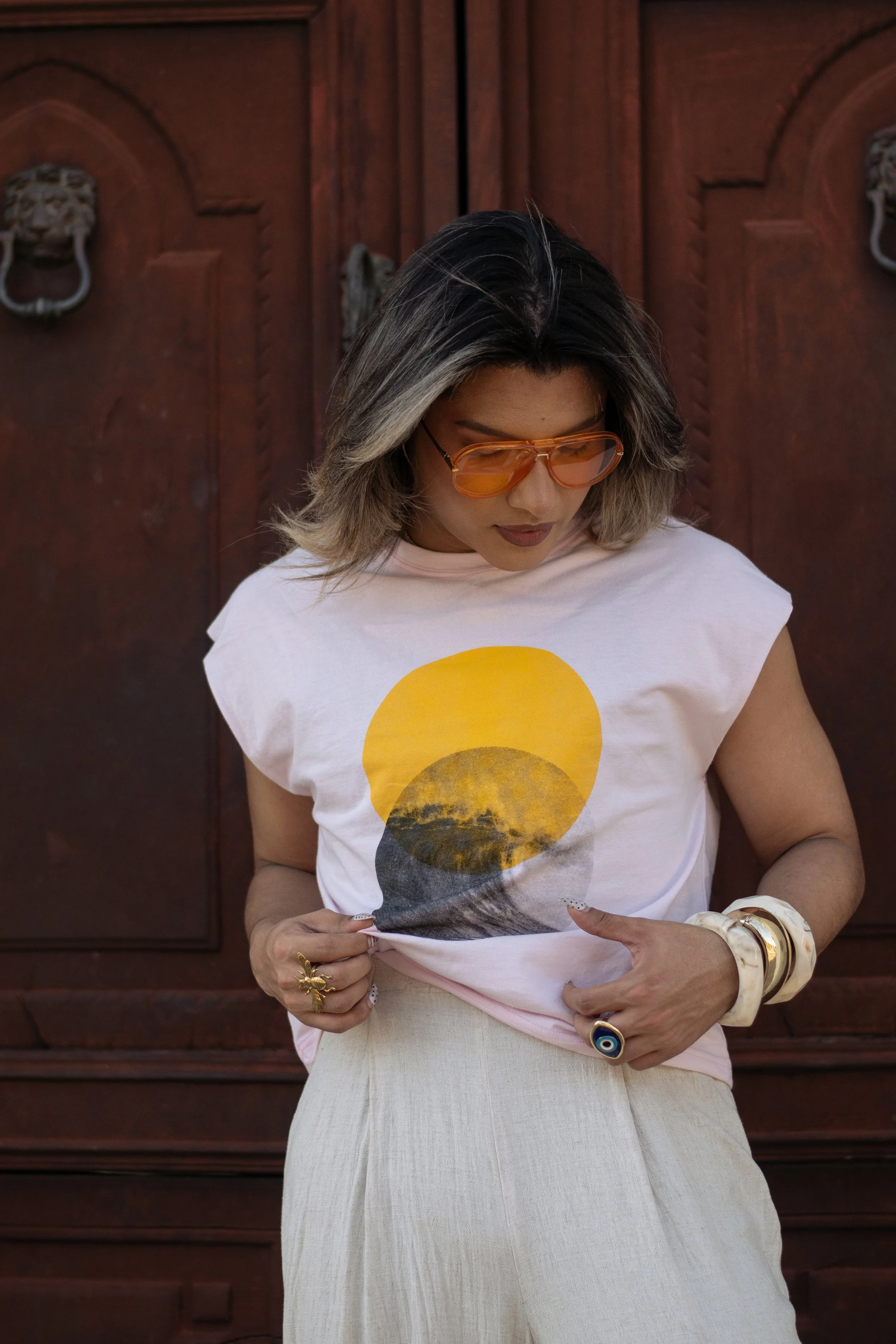 Woman with shoulder-length gray and black hair, wearing orange sunglasses, a light pink T-shirt with a sunset graphic, beige skirt, and various jewelry, standing in front of a dark wooden door.