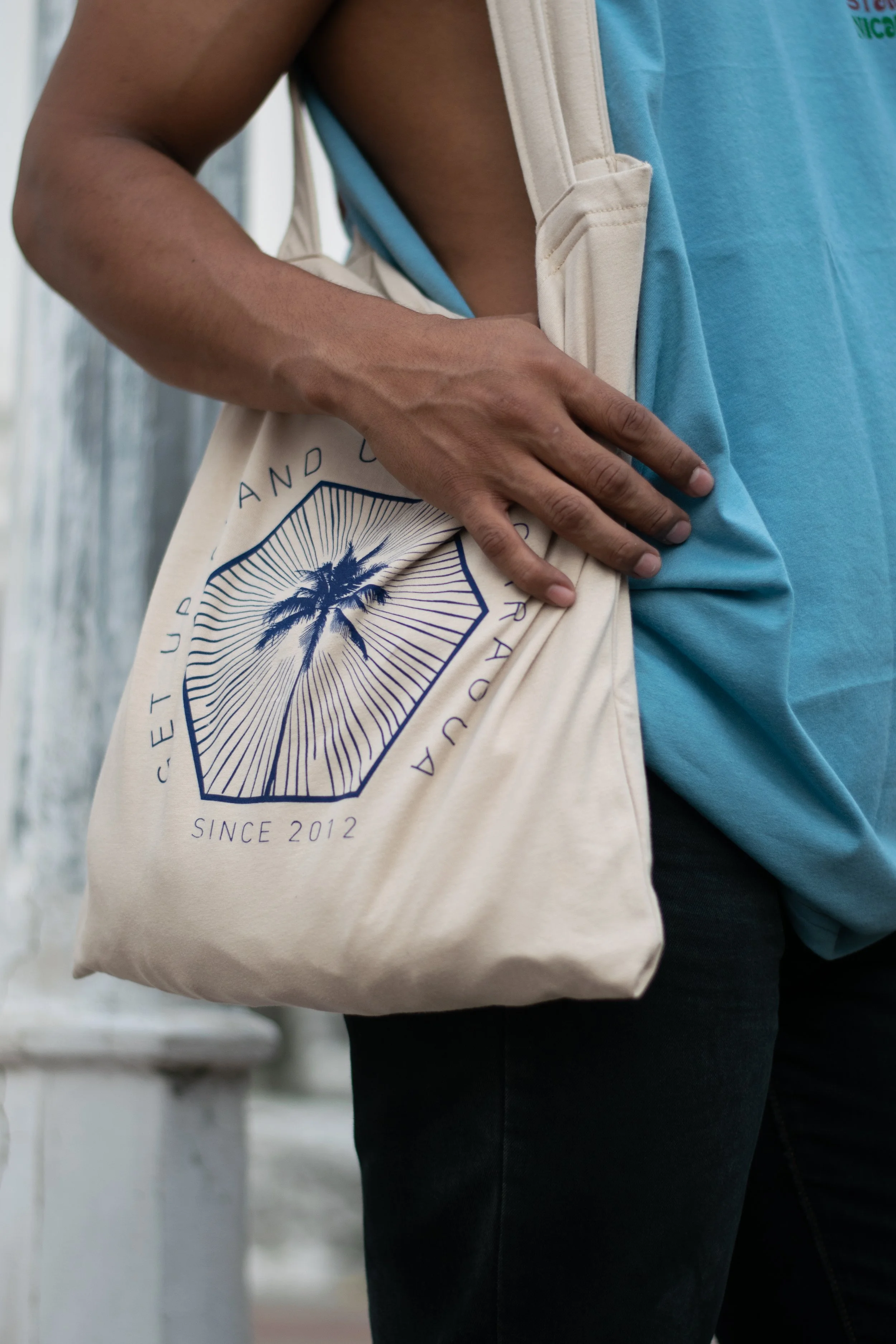 A person is holding a beige tote bag with a palm tree logo and text that reads "Since 2012". They are wearing a tan vest over a blue shirt, and their hand rests on their waist. The background appears to be a white painted wooden wall.