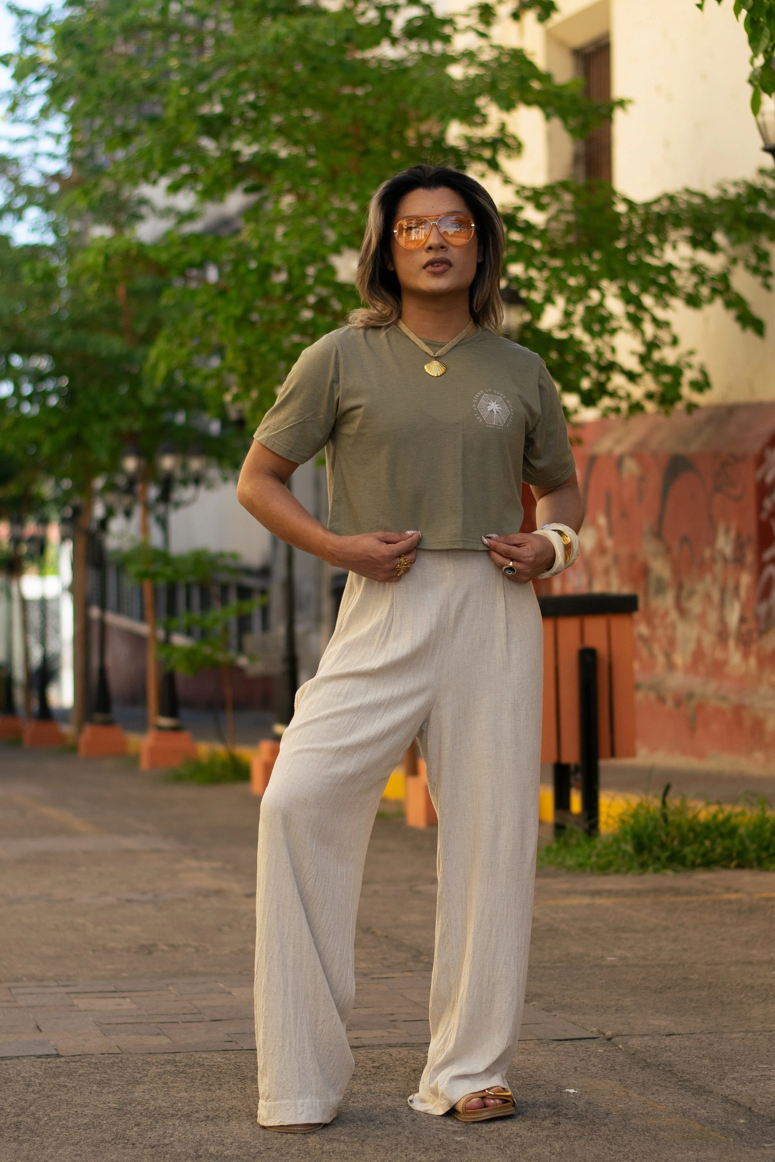 A woman with shoulder-length dark hair and sunglasses standing outdoors on a paved path with trees and a building in the background, wearing a olive green crop top with a small circular logo on the chest, wide beige pants, gold jewelry, and tan sandals.