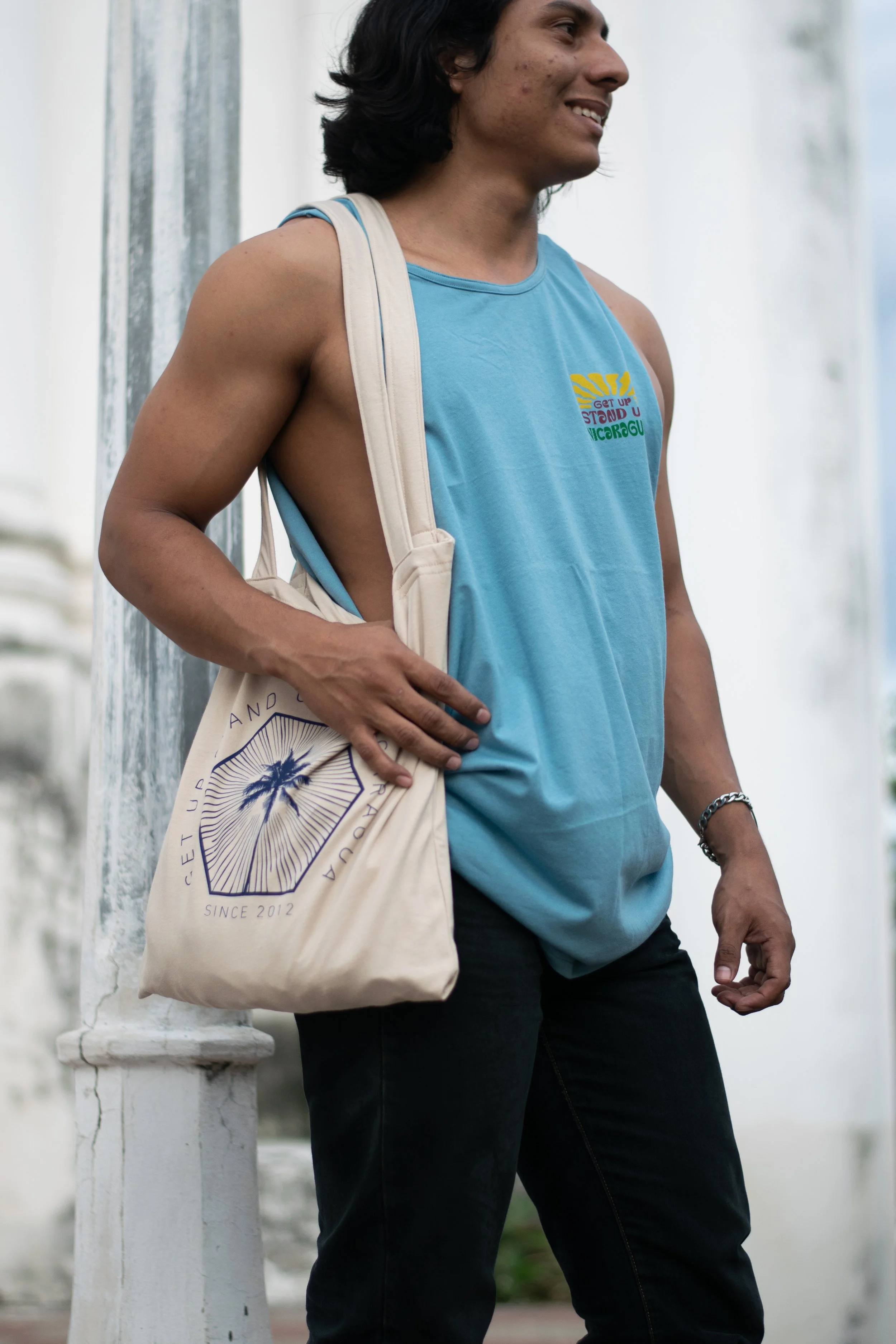 A young man casually standing outdoors next to a white column, wearing a blue sleeveless shirt, black pants, and a silver bracelet, carrying a tote bag on his shoulder, with a smile on his face and dark hair.