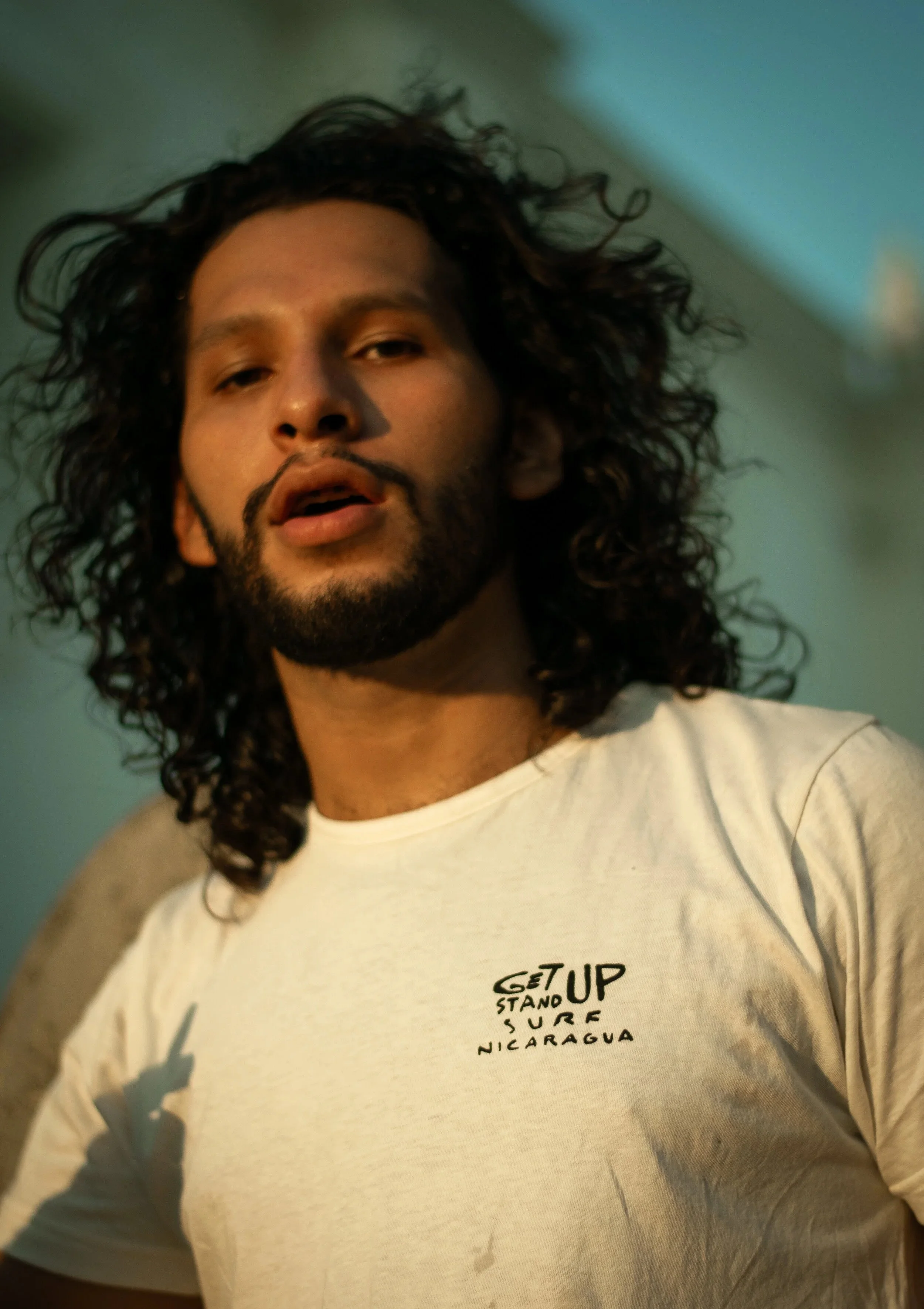 Close-up of a man with long, curly dark hair and a beard, wearing a white sweatshirt with text about Nicaragua, standing indoors against a plain wall.