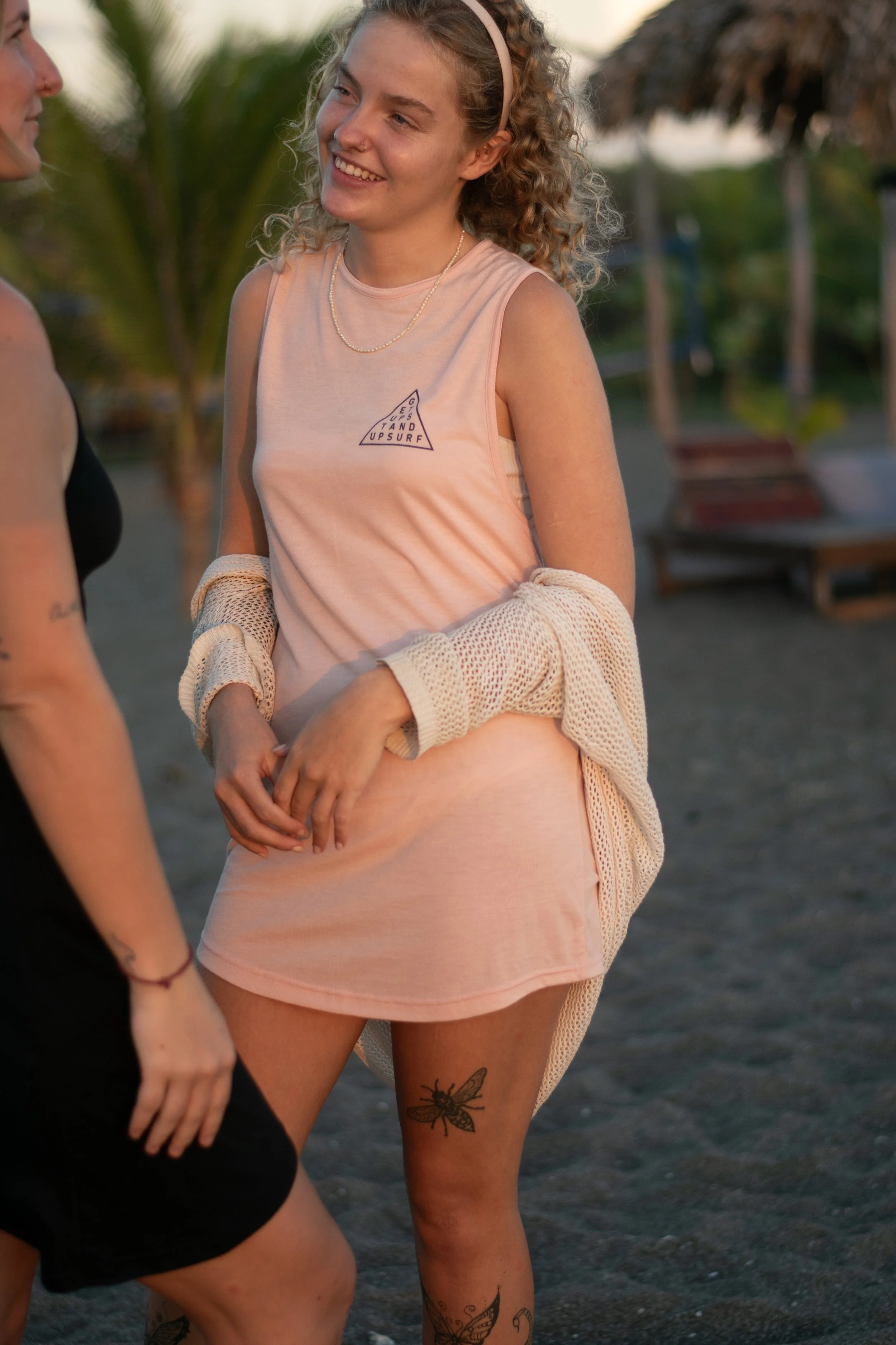 Two young women standing and talking outdoors at sunset, with palm trees and benches in the background. One woman has curly hair, a headband, and a dragonfly tattoo on her thigh, and is smiling while wearing a sleeveless shirt and a crochet cardigan.