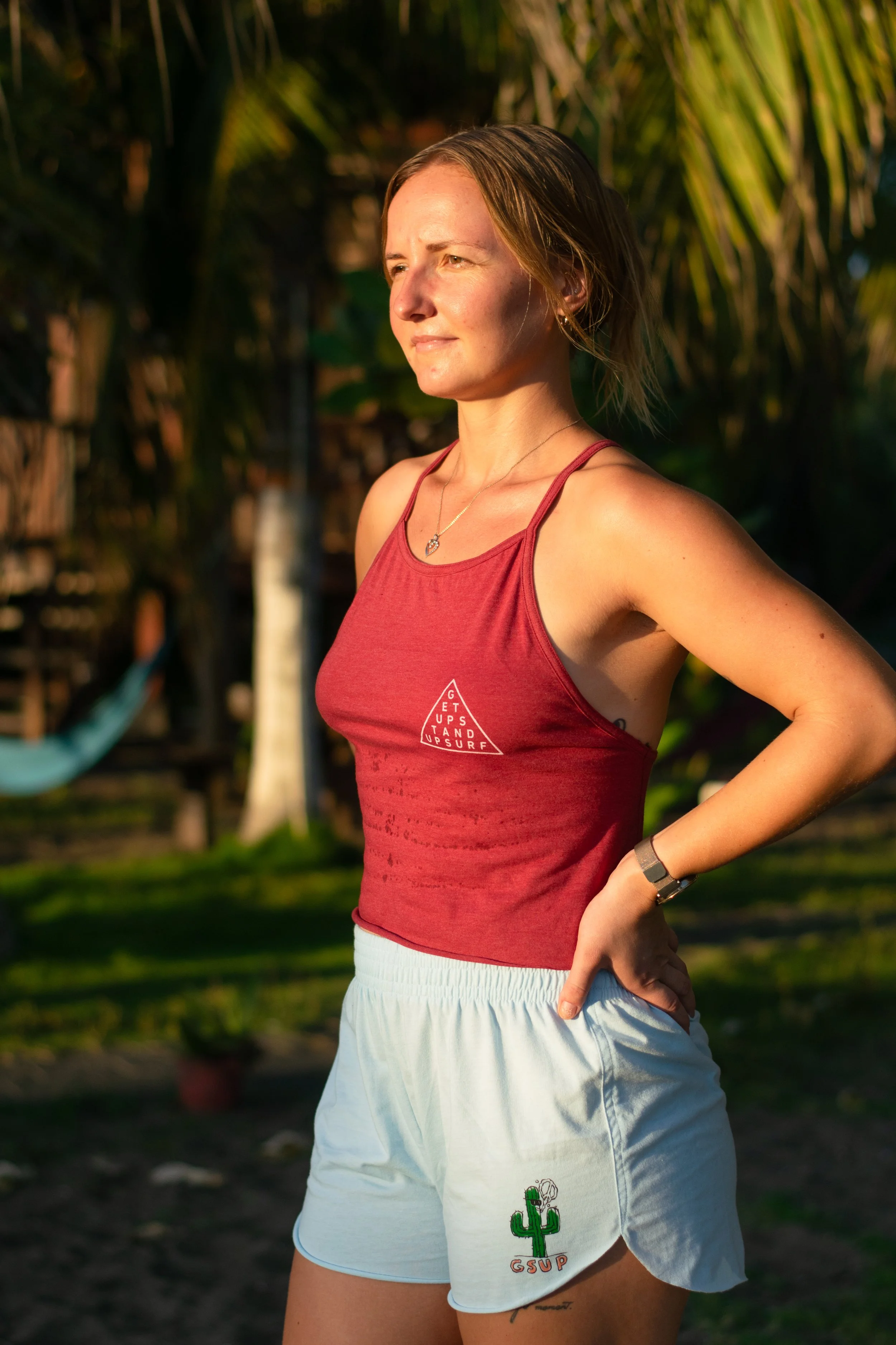A woman stands outdoors in warm sunlight, wearing a red tank top and white shorts with a cactus graphic and the letters 'GSSUP'. She has short, light brown hair, a necklace, a smartwatch, and a tattoo on her thigh. There are trees and a hammock in the background.