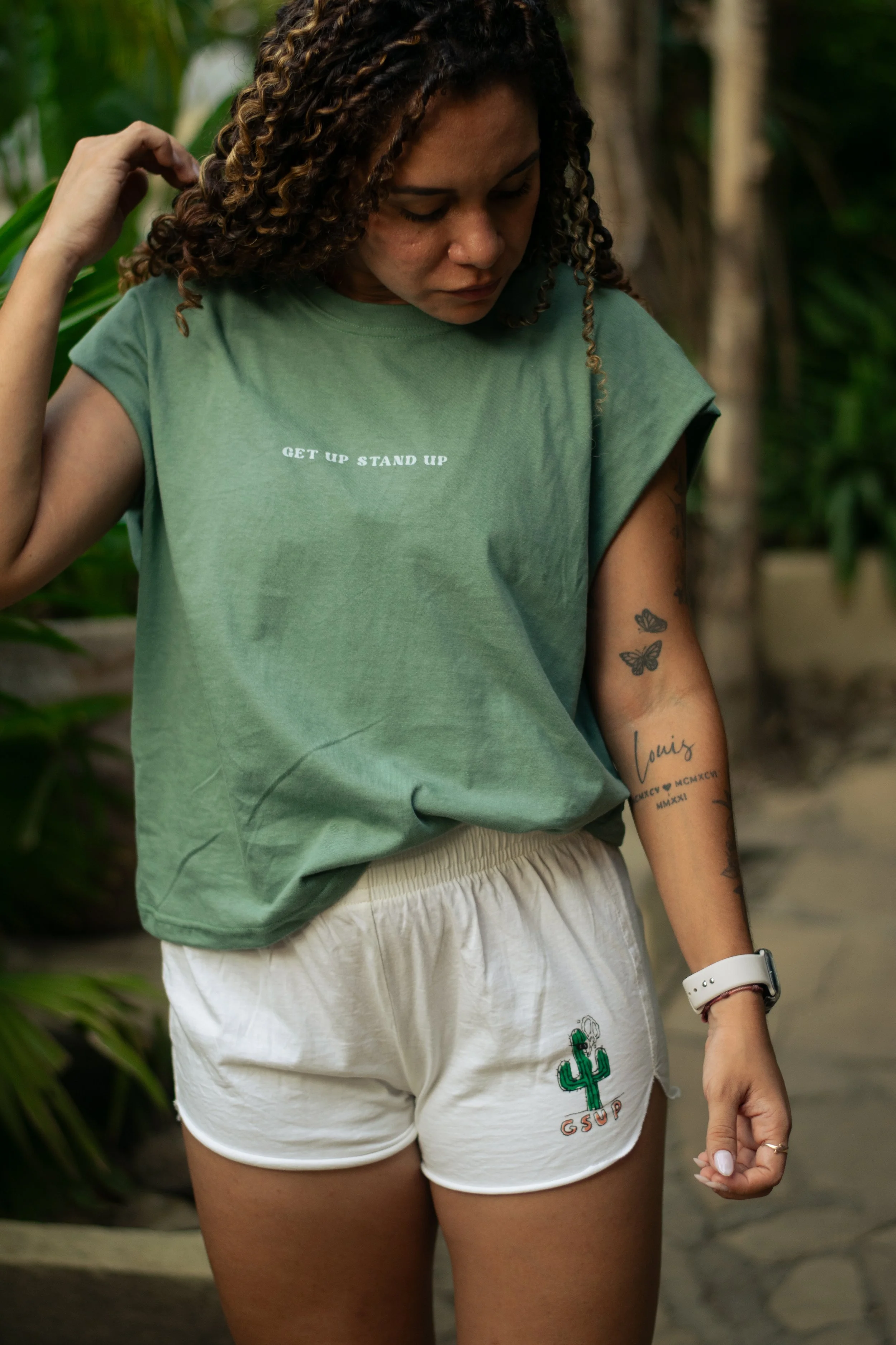 A woman with curly hair wearing a green T-shirt with the words 'GET UP STAND UP' on it, and white shorts with a cactus design and the initials 'GSUP'. She appears to be adjusting her hair or touching her head, with tattoos on her arm, in a lush green outdoor setting.