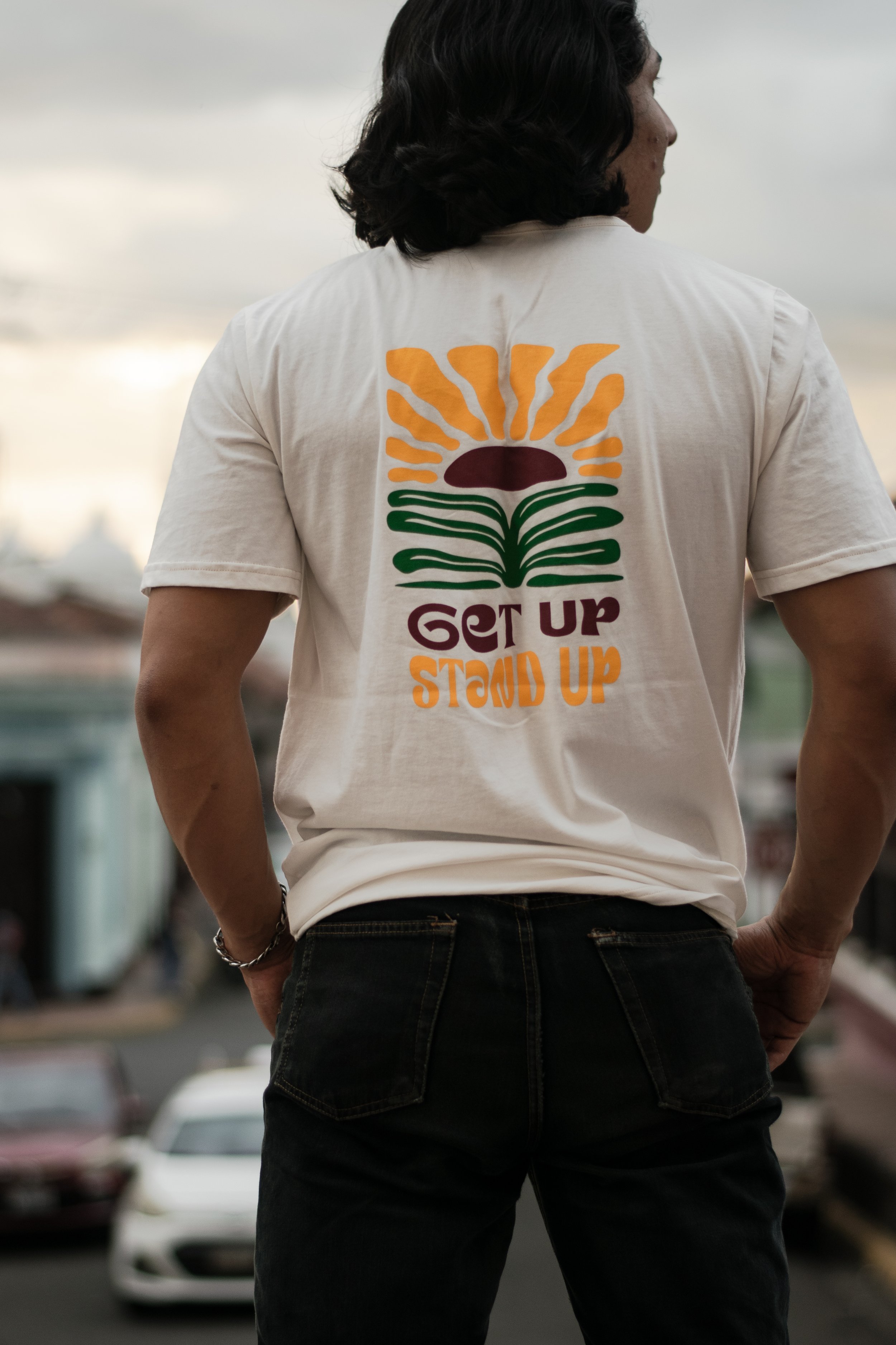 Person with dark, curly hair wearing a white T-shirt with a colorful graphic and text that reads 'GET UP STAND UP,' standing outdoors near parked cars.