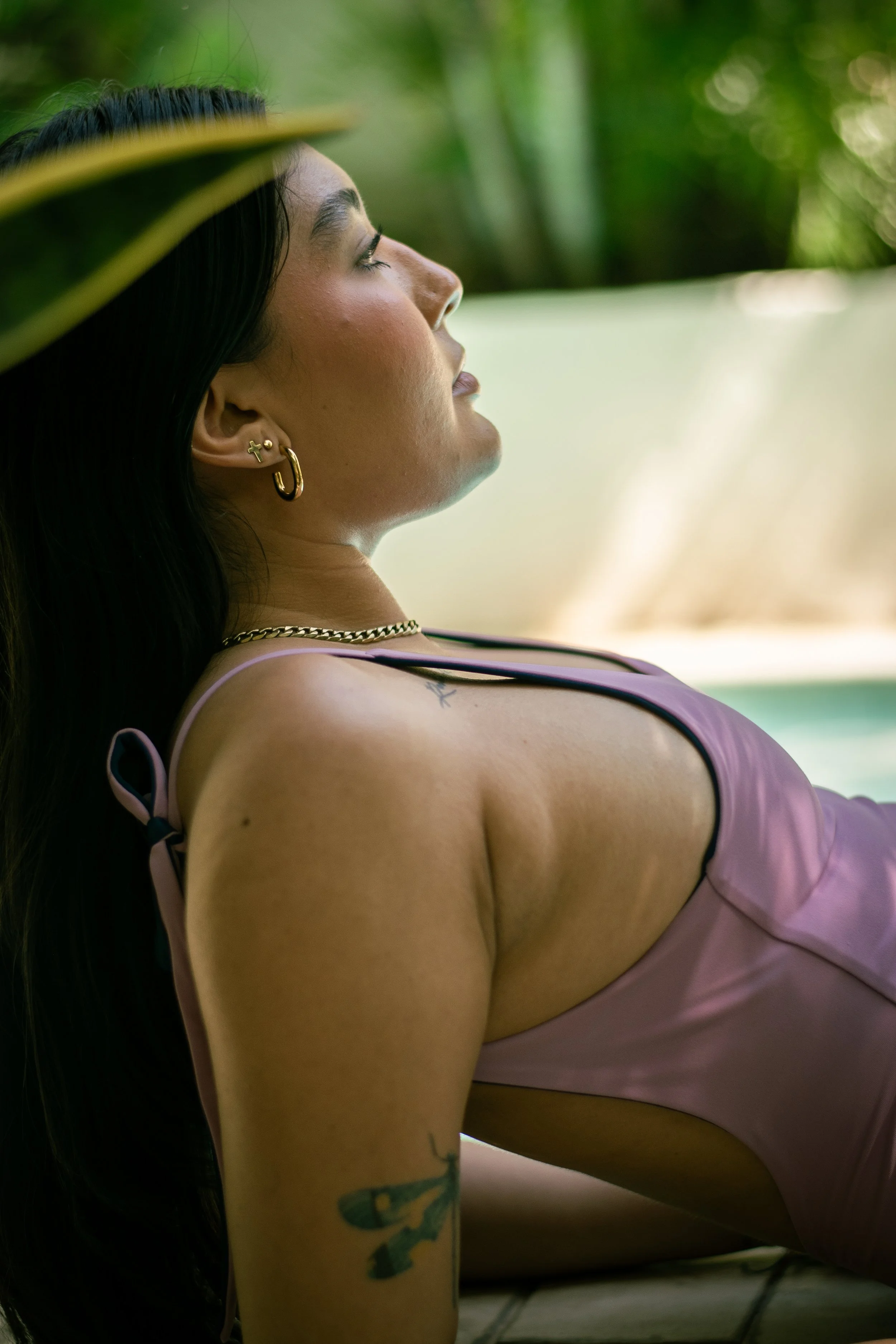 Close-up side profile of a woman with long dark hair, gold earrings, and a chain necklace, wearing a pink satin top, leaning back with greenery in the background.