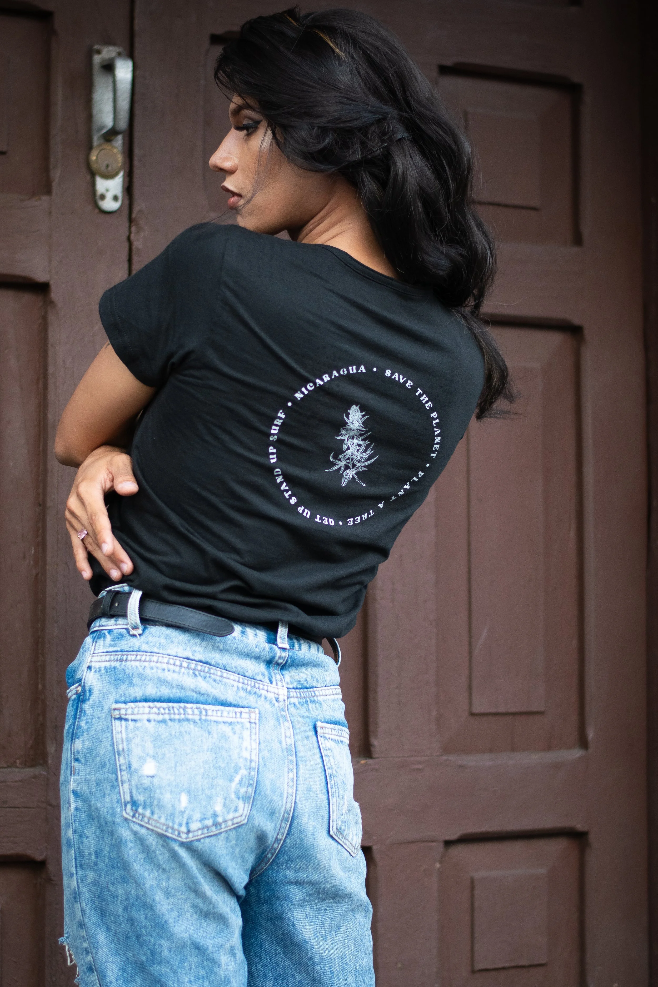 A woman with dark, wavy hair wearing a black t-shirt and light blue jeans standing in front of a brown wooden door.