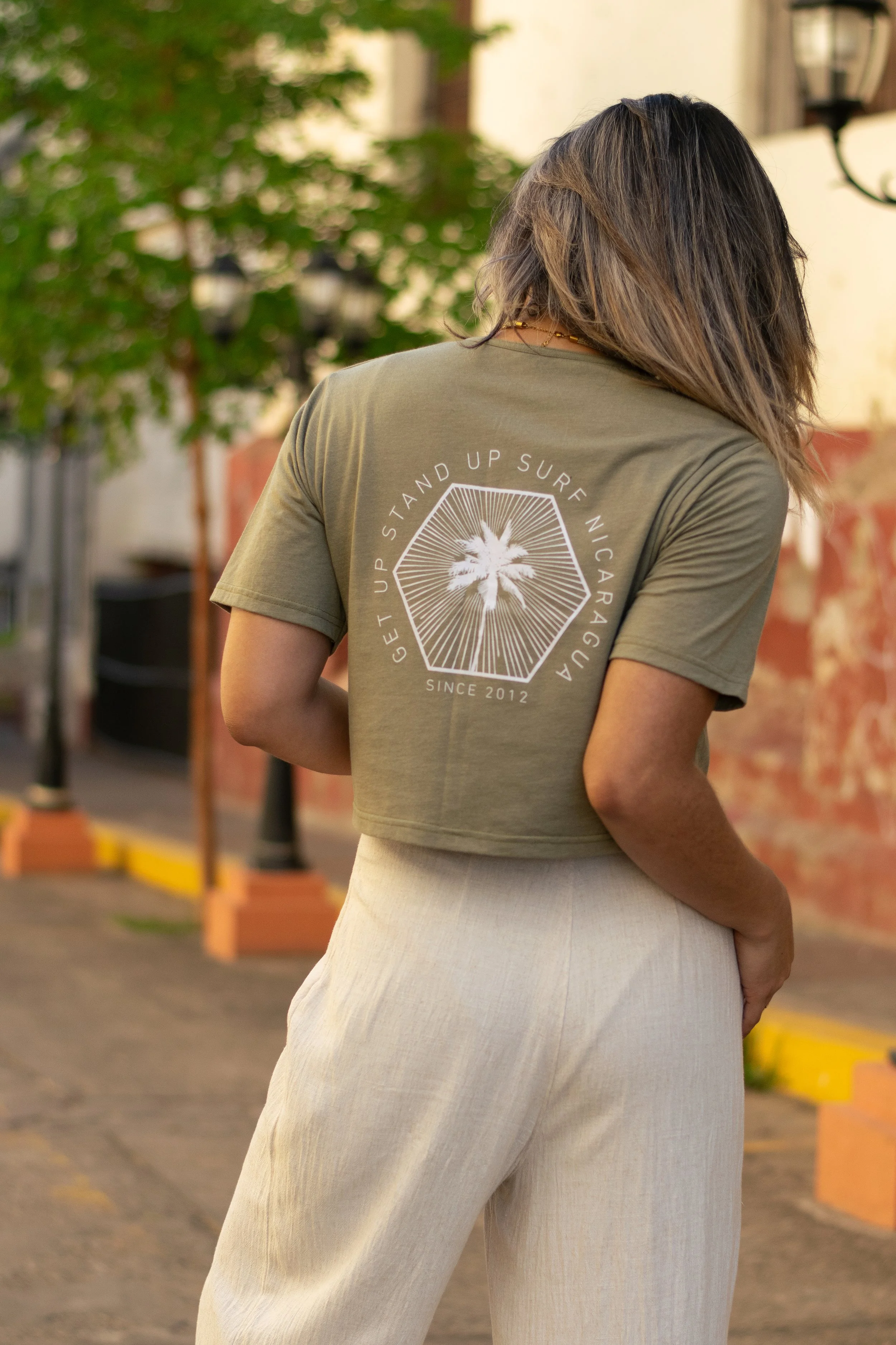 Back view of a woman with shoulder-length wavy hair wearing a green T-shirt with a graphic and text on the back, and beige pants, standing outdoors in an urban area with trees, a street lamp, and a brick wall in the background.