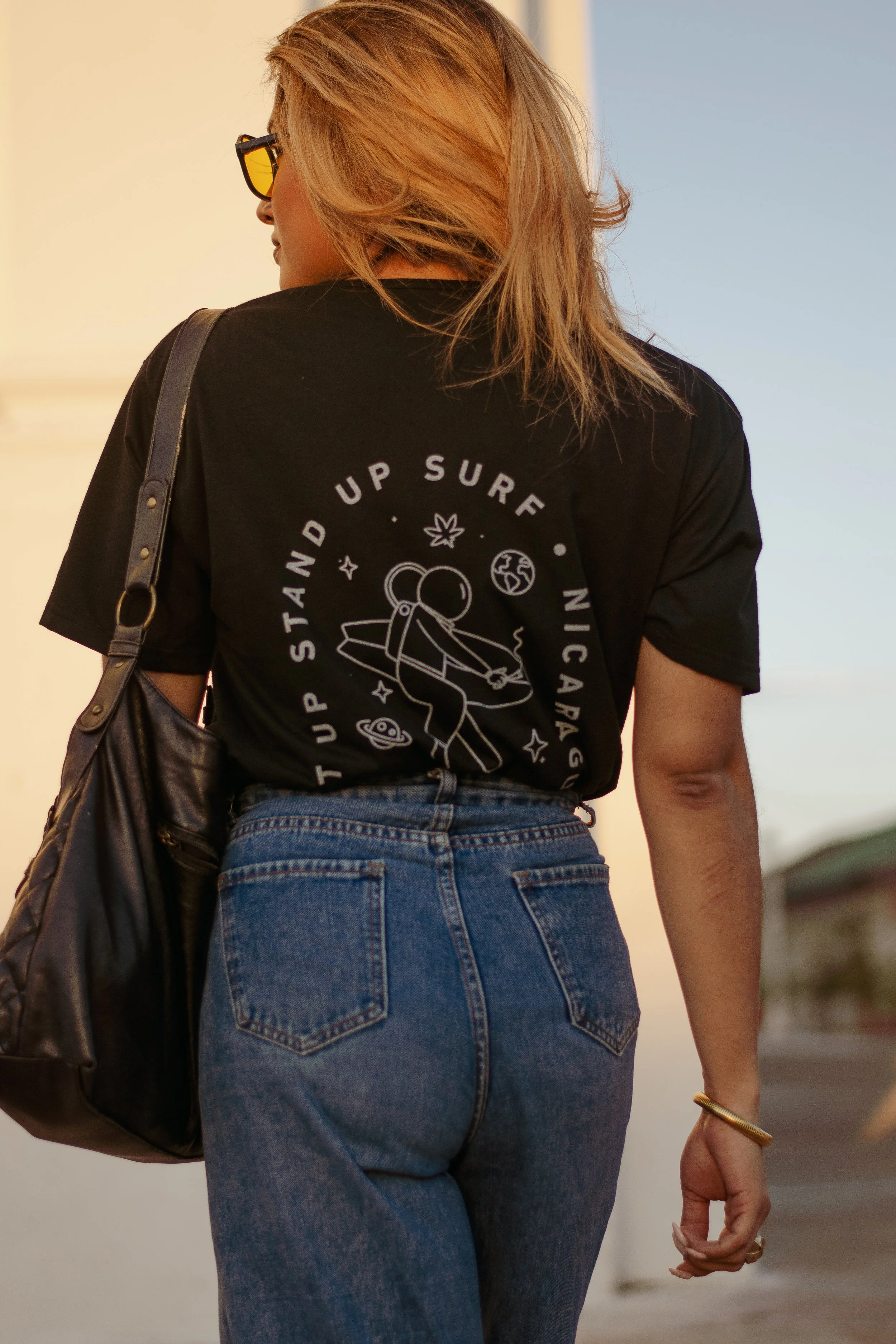 A person with light brown hair wearing sunglasses, a black T-shirt with a space-themed design, and blue jeans, walking outdoors. The T-shirt has white print with space-related graphics and text.