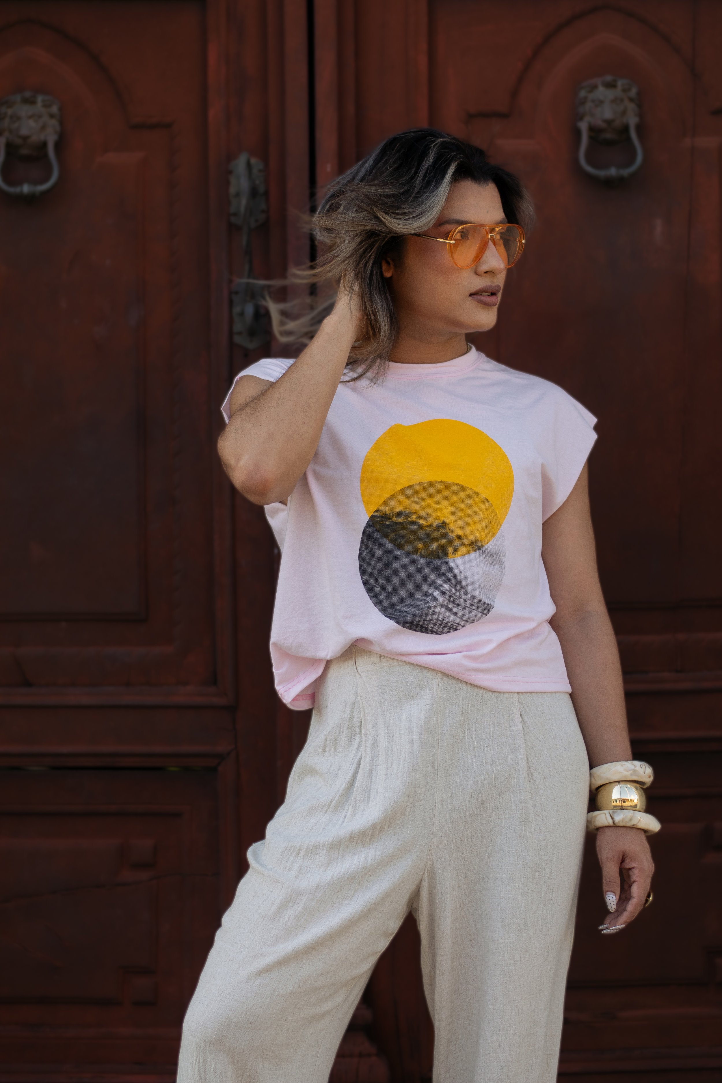 Woman wearing orange sunglasses, a white T-shirt with a sun and mountain graphic, and cream-colored pants, standing in front of wooden doors with lion head knockers.