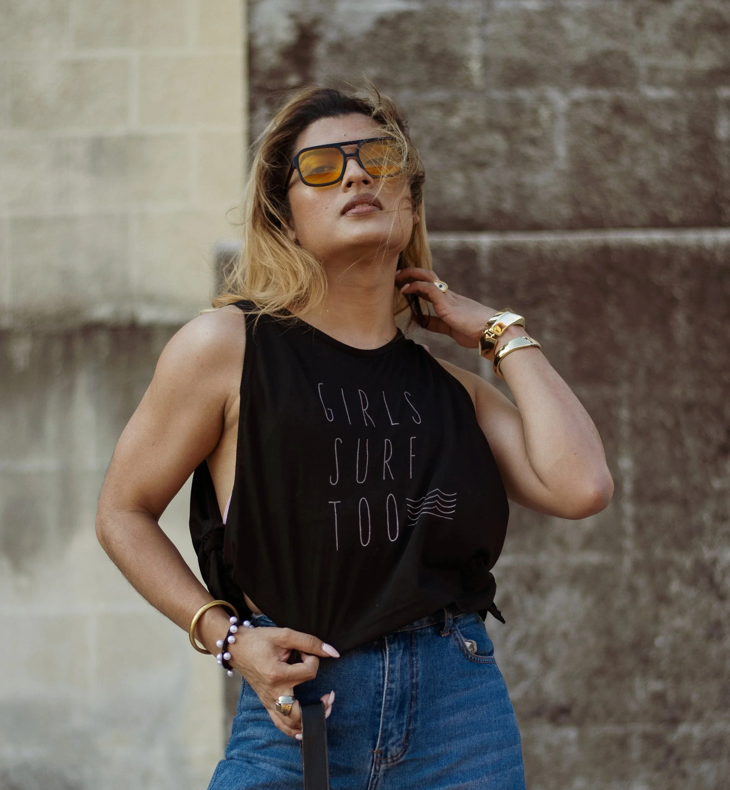 A woman with blonde hair wearing large yellow sunglasses, a black sleeveless top with white text, and blue jeans, standing against a stone wall. She is accessorized with gold jewelry and bracelets, and has her hand resting on her neck.
