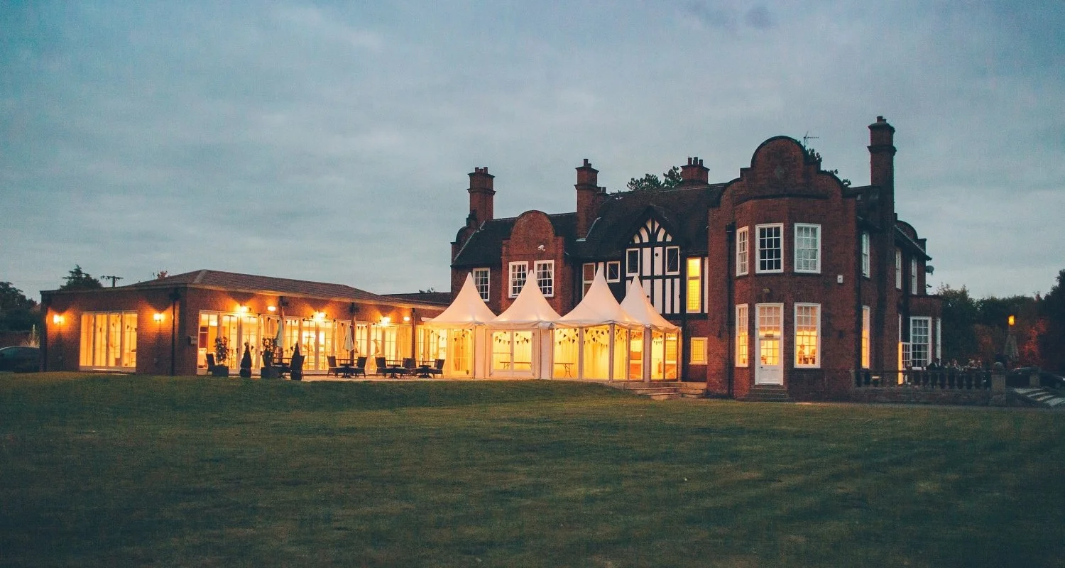 A large brick house illuminated with warm lights, featuring a glass extension and white tent structures in the garden during dusk.