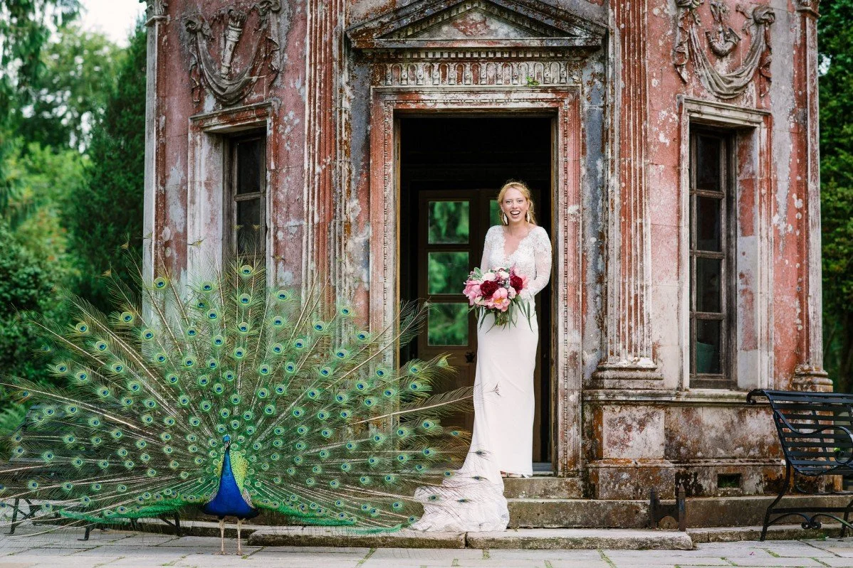 A bride in a white wedding dress holding a bouquet of pink flowers, standing at a doorway of an old, weathered building, with a peacock displaying its feathers in front of her.