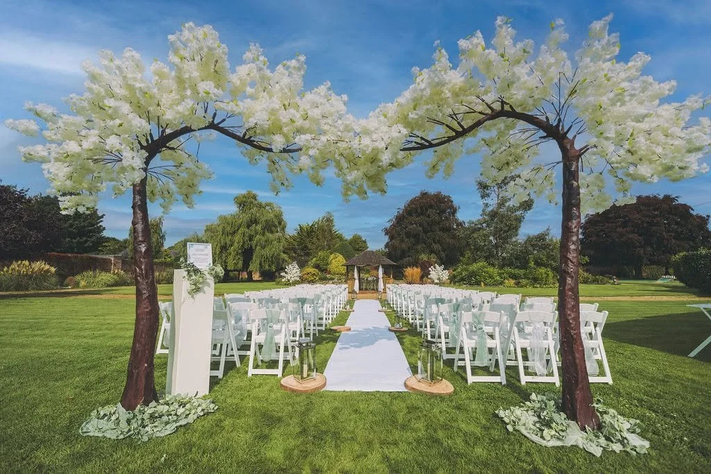 Outdoor wedding ceremony setup with white chairs arranged on either side of a white aisle runner, under decorative trees with white blossoms, with a gazebo in the background on a sunny day with blue sky.