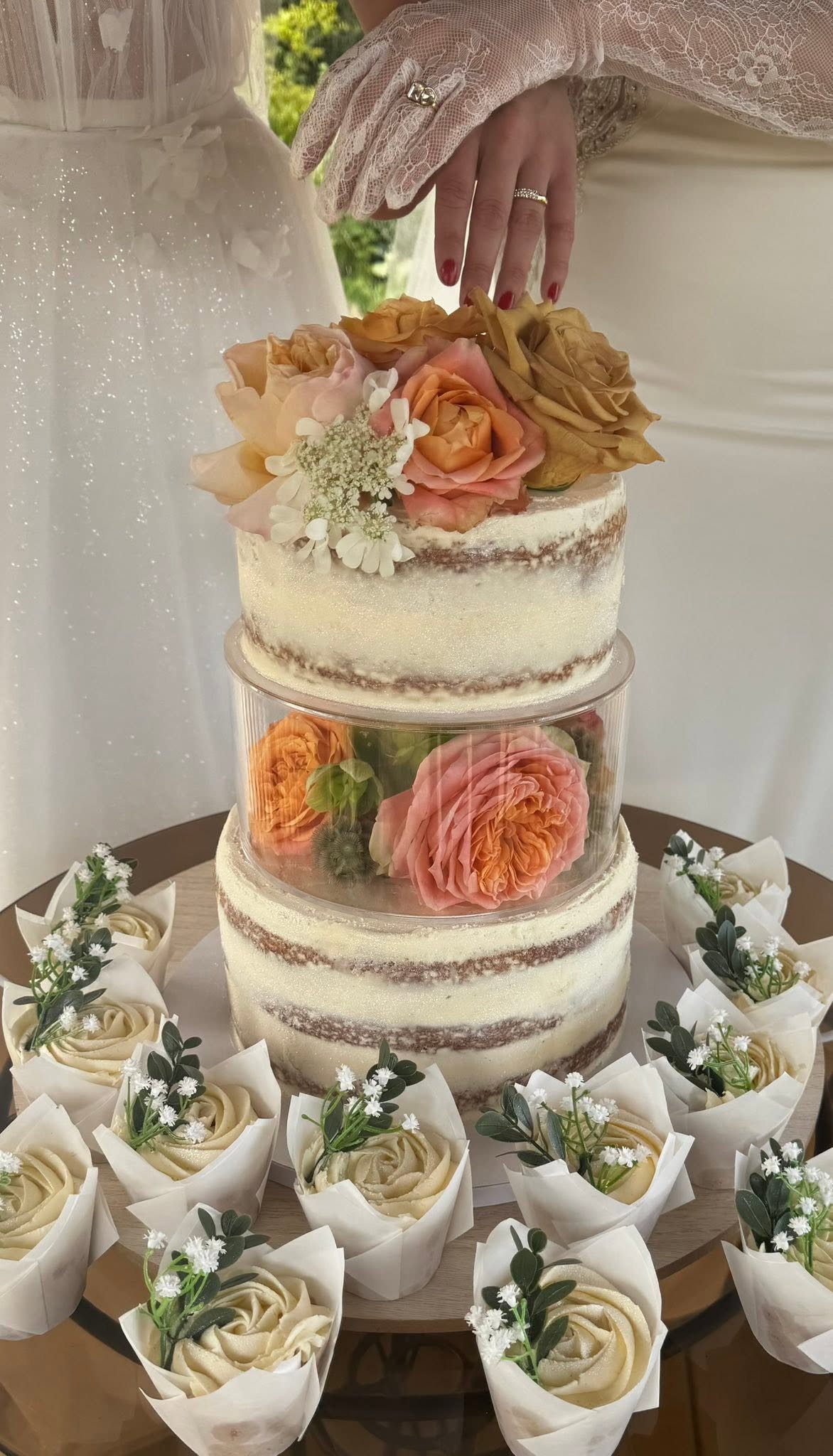 Wedding cake with three tiers decorated with peach, pink, and brown roses, topped with a floral arrangement. Surrounding the cake are small white cupcakes with cream roses and greenery in paper wrappers, arranged on a round tray.