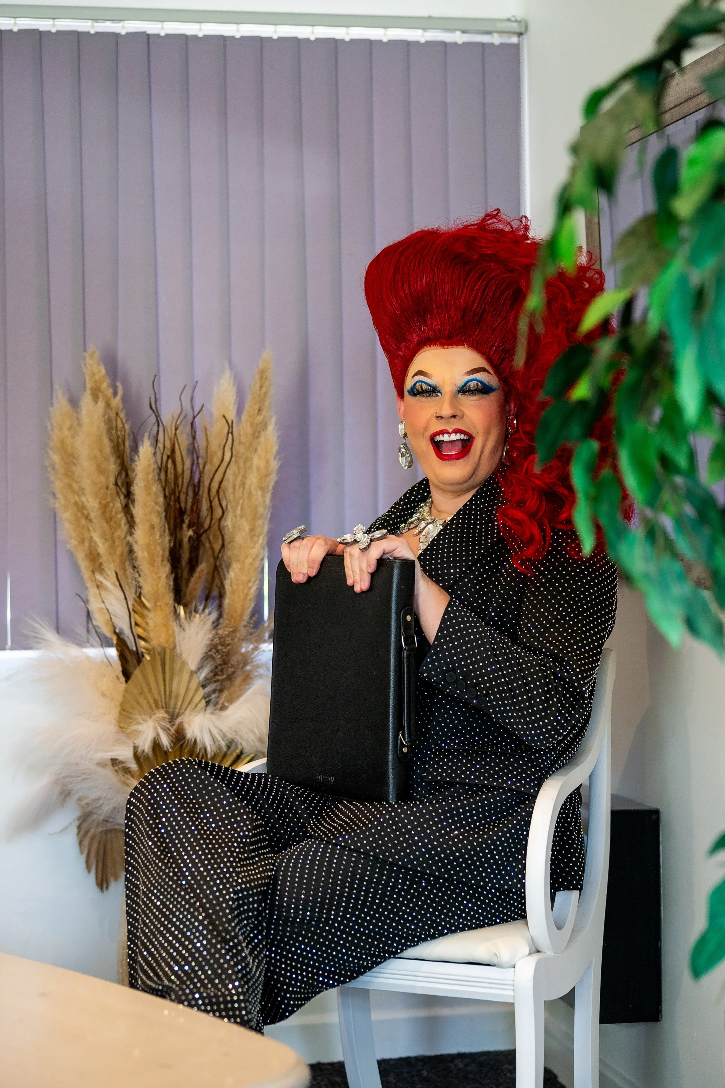 A person with bright red hair styled high, wearing bold makeup and polka dot clothing, smiling and holding a briefcase while sitting on a white chair in an indoor setting. There are decorative pampas grass and green plant leaves in the background, with vertical blinds partially visible.