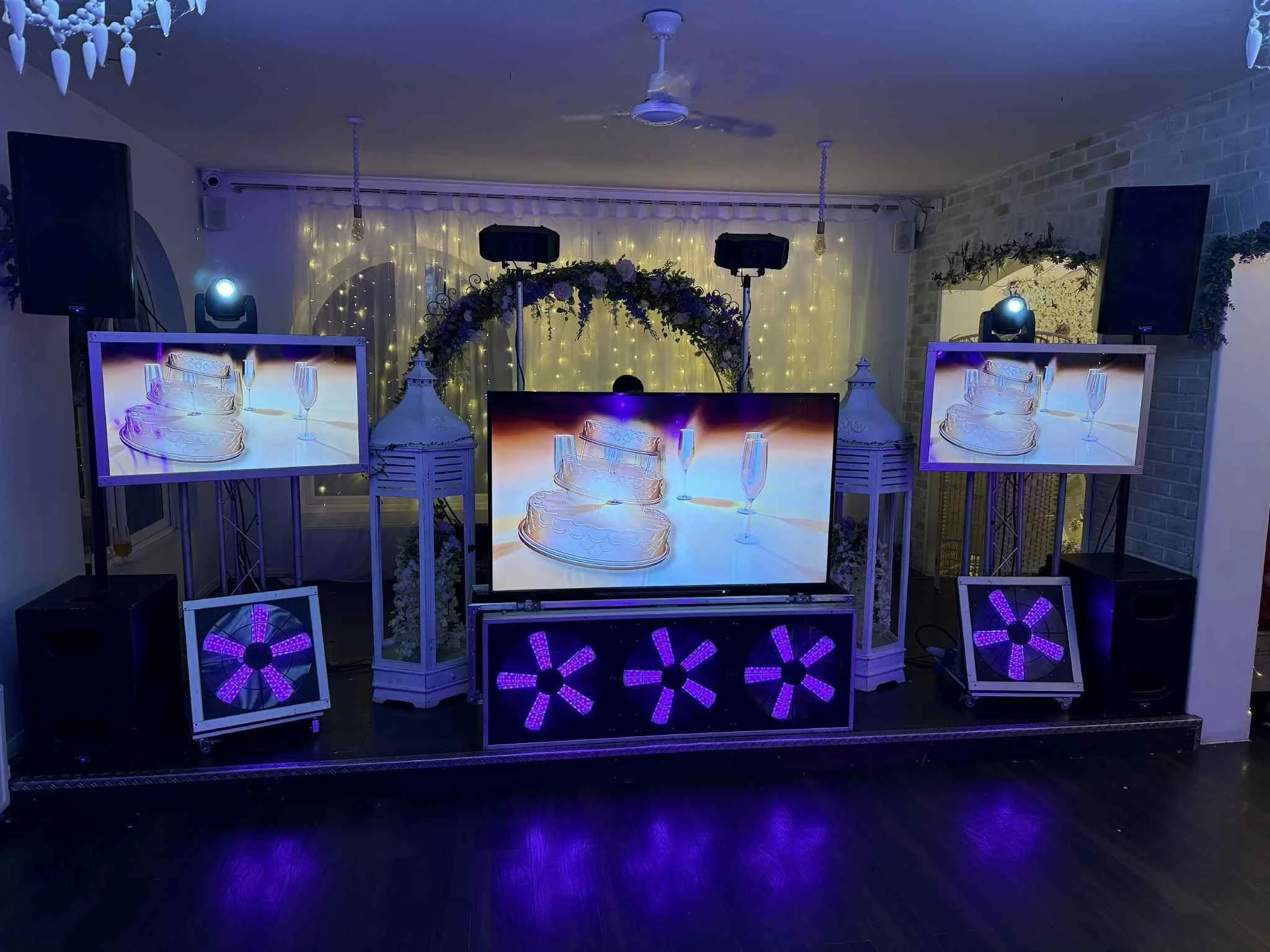 Decorative party setup with LED screens showing champagne glasses and cake, floral arch, black speakers, and purple lighting.