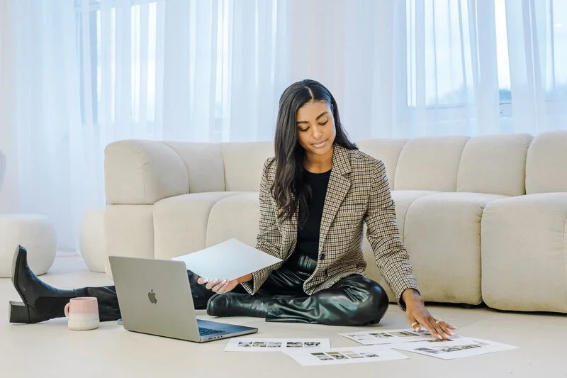 Fashionably dressed woman sitting on the floor in front of a beige sofa, working with printed materials, a laptop, and a cup in a bright living room.