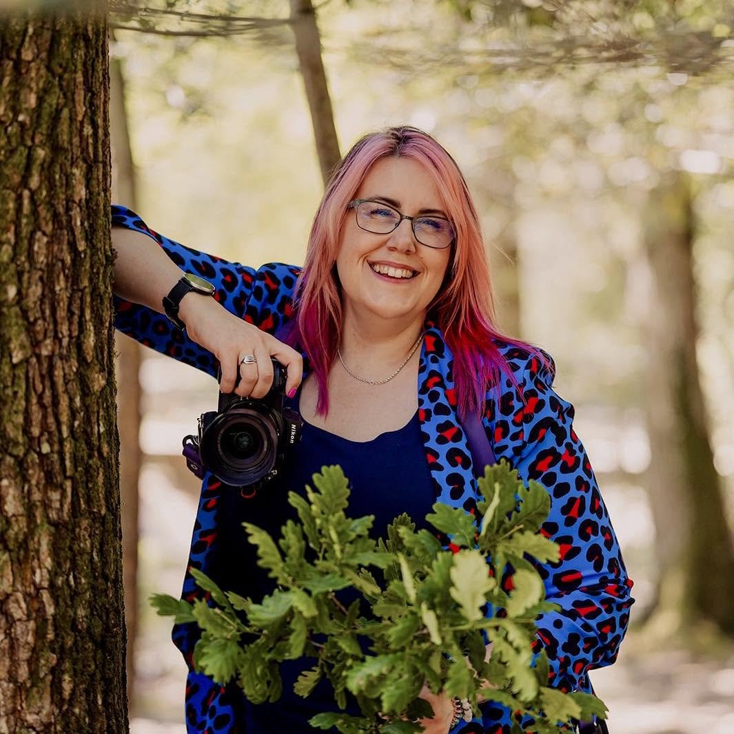 Woman with pink hair, glasses, and a colorful animal print jacket smiling outdoors with trees in the background, holding a camera in one hand and a plant in the other.